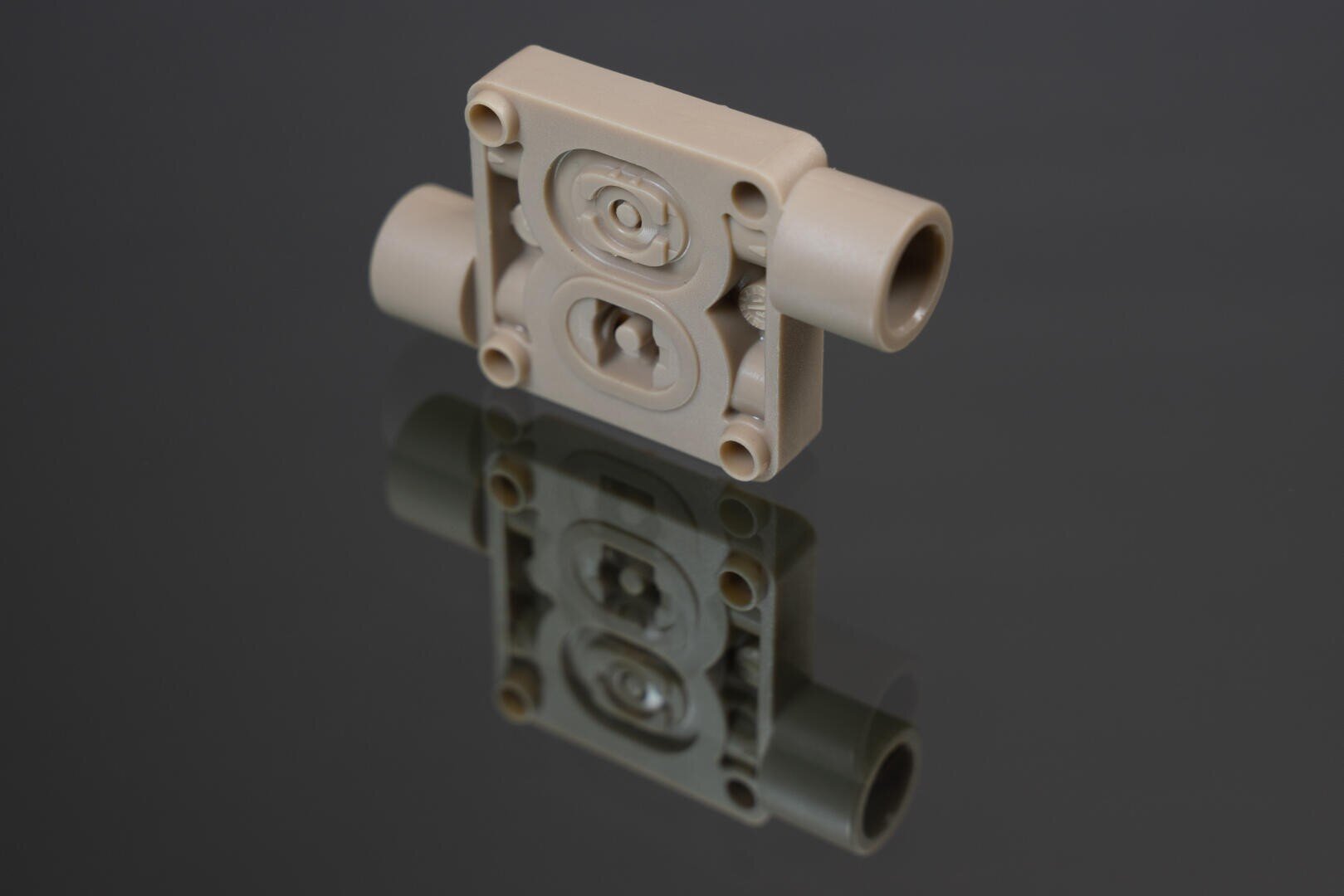 Ein beigefarbenes mechanisches Kunststoffteil mit zylindrischen Anschlüssen auf jeder Seite wird auf eine reflektierende dunkle Oberfläche gelegt, so dass es sich darunter spiegelt. Das Teil hat eingekerbte Muster und vier kleine Löcher an den Ecken.