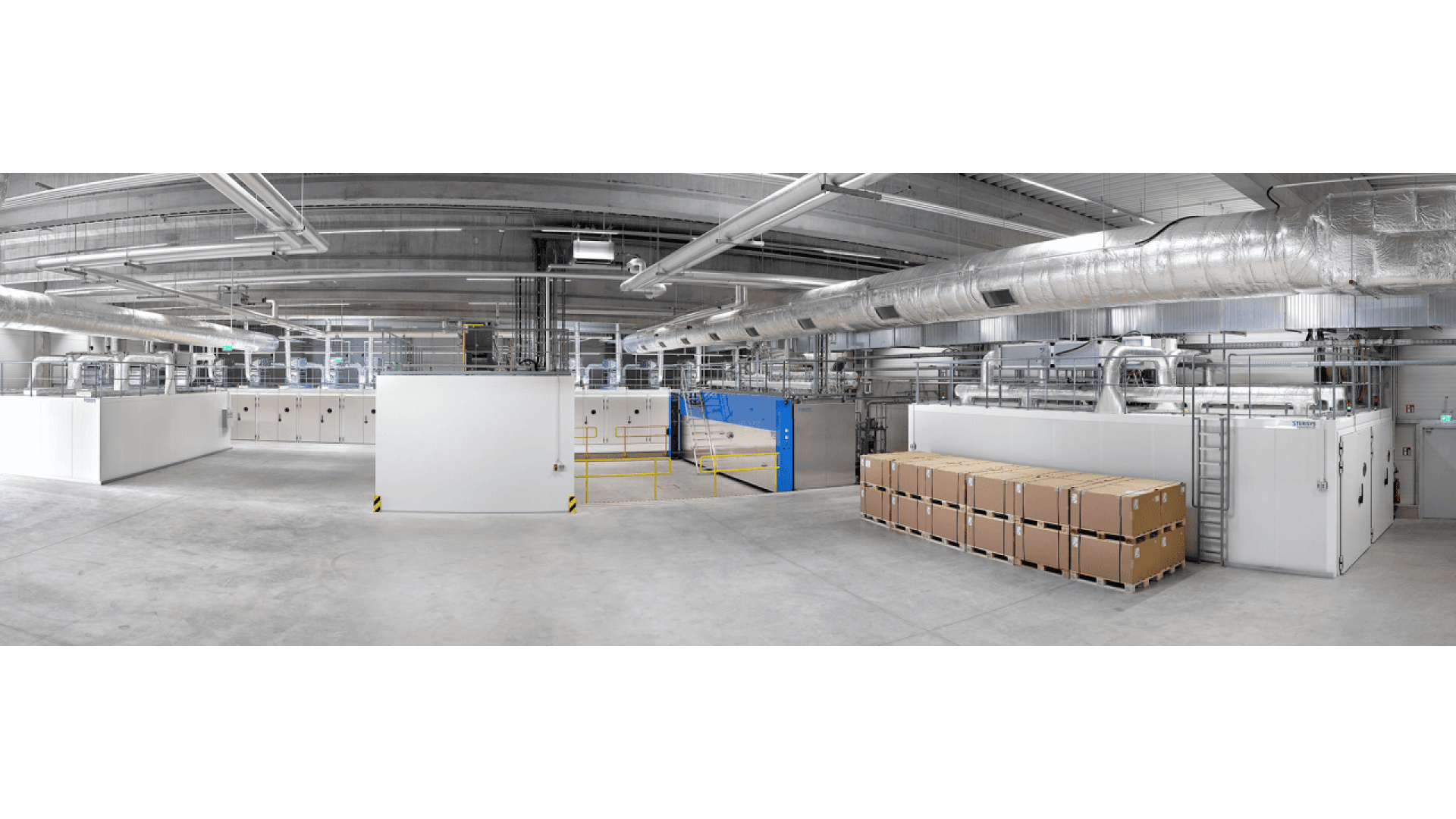 A spacious industrial facility with exposed ductwork, white walls, and concrete floors. Stacks of sealed cardboard boxes are arranged along the right side, with machinery and equipment visible in the background.