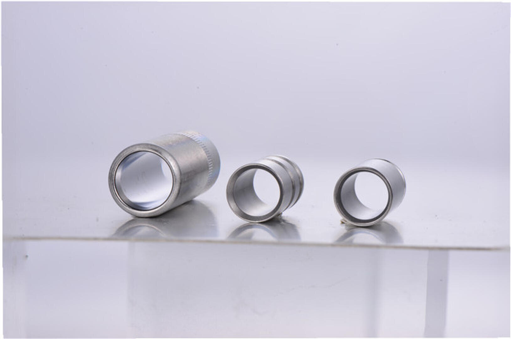 Drei unterschiedlich große zylindrische Metallringe werden nebeneinander auf einer klaren, reflektierenden Oberfläche vor einem hellen Hintergrund platziert.
