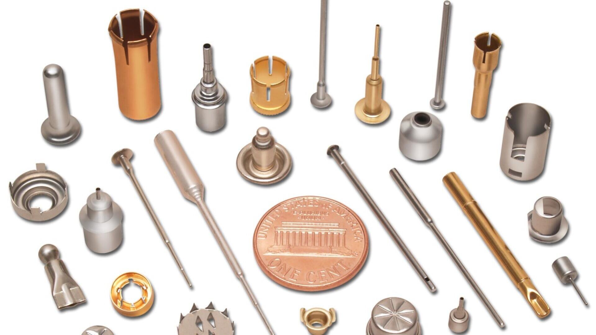 Various small, metallic precision parts of different shapes and finishes are arranged in a circle around a U.S. penny, highlighting their tiny size and detailed craftsmanship.