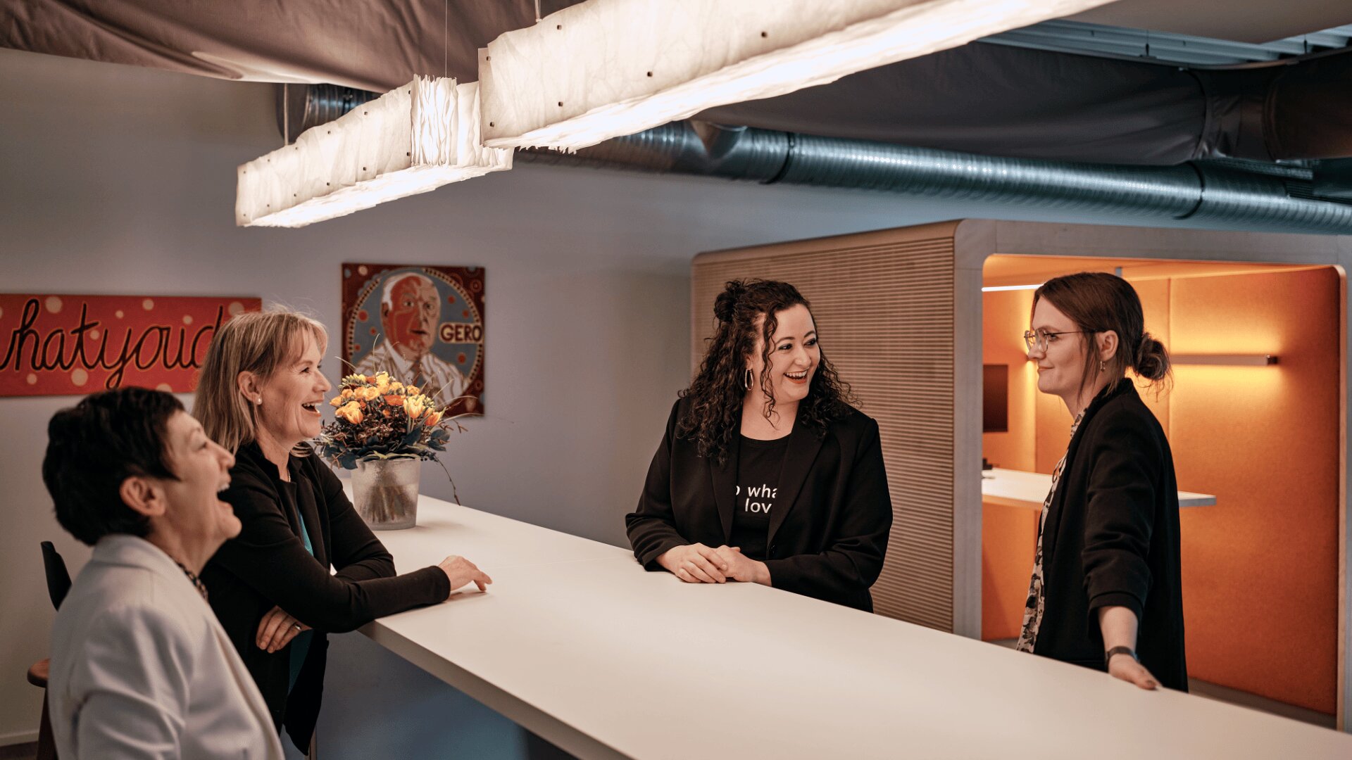 Vier Frauen stehen lächelnd und unterhalten sich um einen hohen weißen Tisch in einem modernen Büro. Hinter ihnen hängt ein Blumenstrauß, ein Kunstwerk an der Wand und eine warme, stimmungsvolle Beleuchtung.