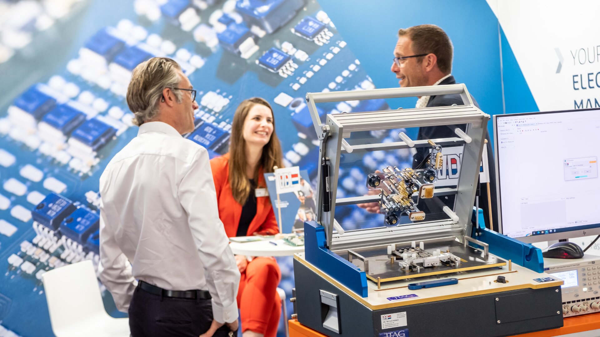 Drei Personen, zwei Männer und eine Frau, unterhalten sich an einem Stand einer Technologiemesse, auf der elektronische Geräte und Leiterplatten ausgestellt sind. Im Vordergrund sind eine Maschine und ein Computeraufbau zu sehen.
