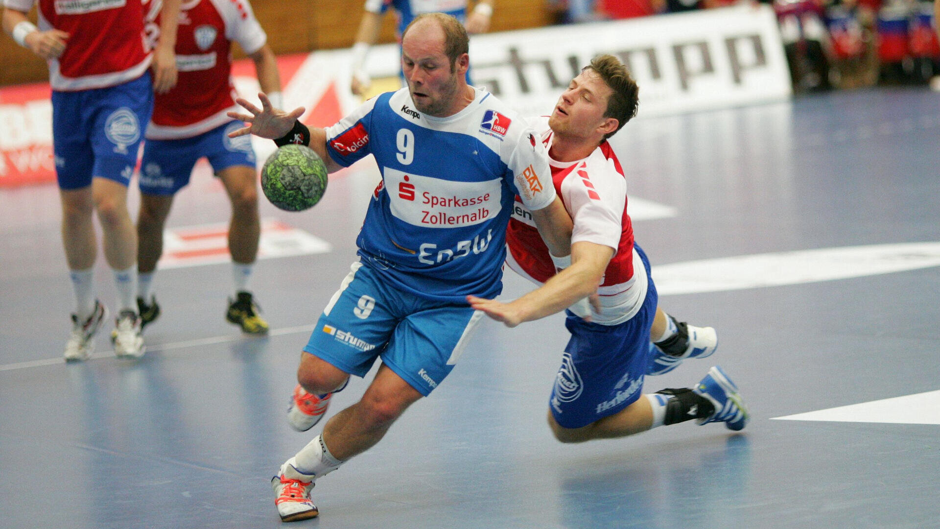 Zwei männliche Handballspieler kämpfen in einer Halle um den Ball. Der Spieler in Blau rennt nach vorn, während der Spieler in Weiß und Rot versucht, ihn zu blocken oder zu tackeln. Im Hintergrund sind weitere Spieler und unscharfe Zuschauer zu sehen.