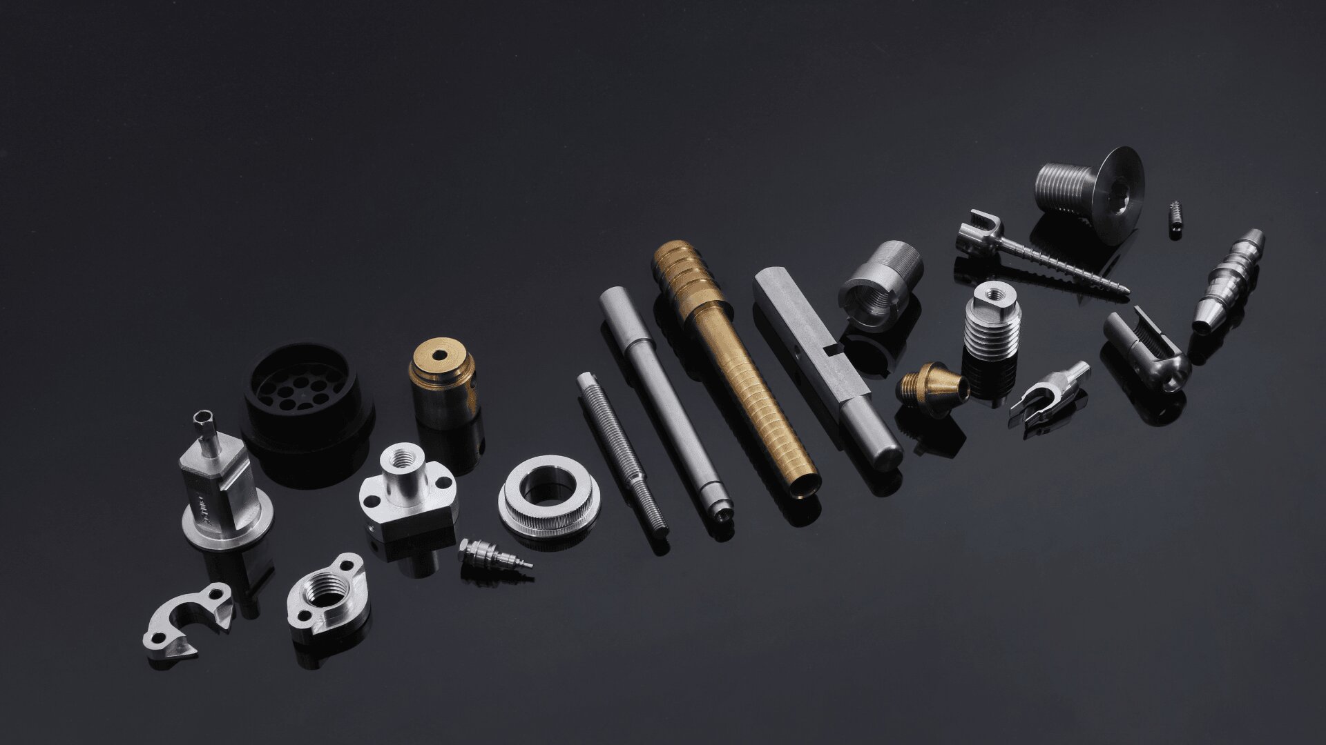 A collection of various precision metal machine parts, including bolts, screws, cylindrical rods, and fittings, arranged neatly on a reflective black surface.