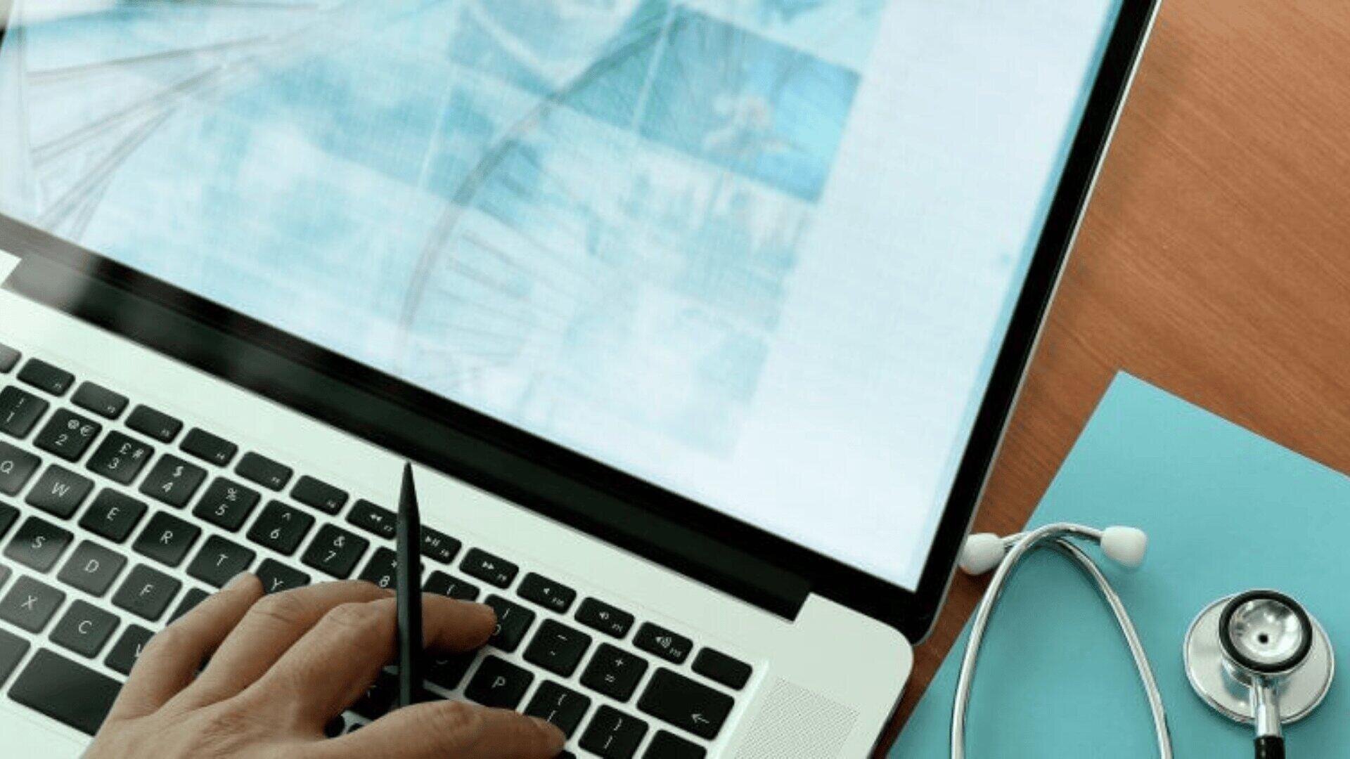 Eine Person benutzt einen Stift auf einer Laptoptastatur, während ein Stethoskop und ein Klemmbrett auf dem hölzernen Schreibtisch in der Nähe liegen, was auf ein medizinisches oder gesundheitliches Umfeld schließen lässt.
