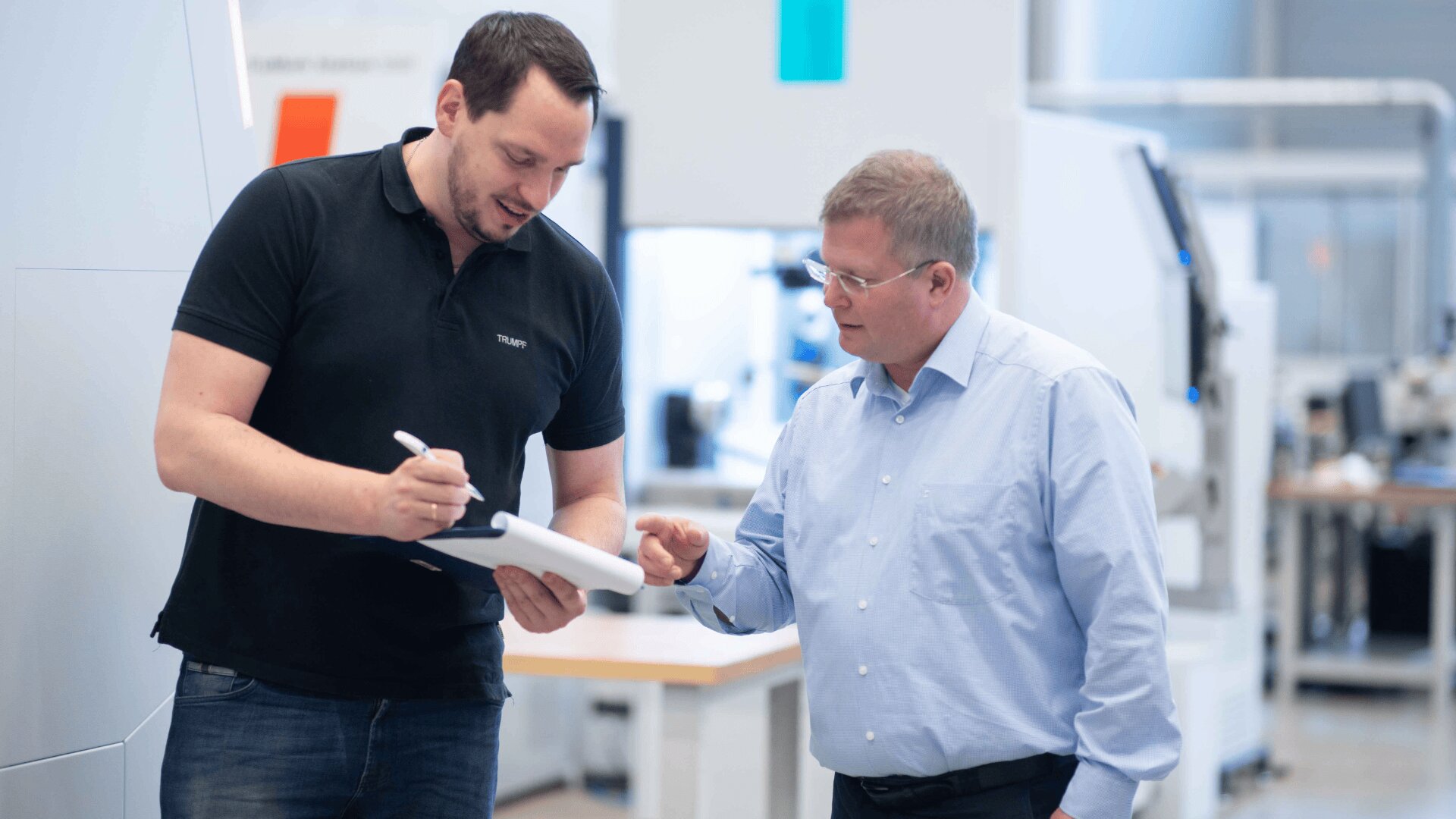Zwei Männer in einer modernen Büro- oder Industrieumgebung besprechen Dokumente auf einem Klemmbrett. Der eine, in einem schwarzen Polohemd, schreibt auf dem Klemmbrett, während der andere, in einem hellblauen Hemd, in Richtung des Papiers gestikuliert.