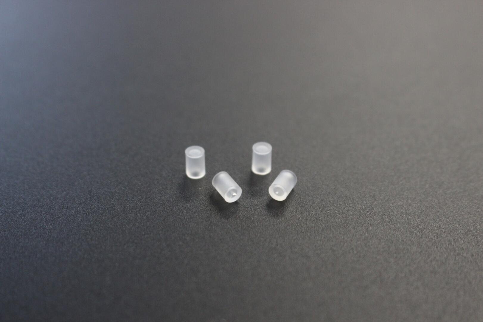 Vier kleine, durchscheinende, zylindrische Kunststoffobjekte, die auf einer glatten, dunkelgrauen Oberfläche angeordnet sind.