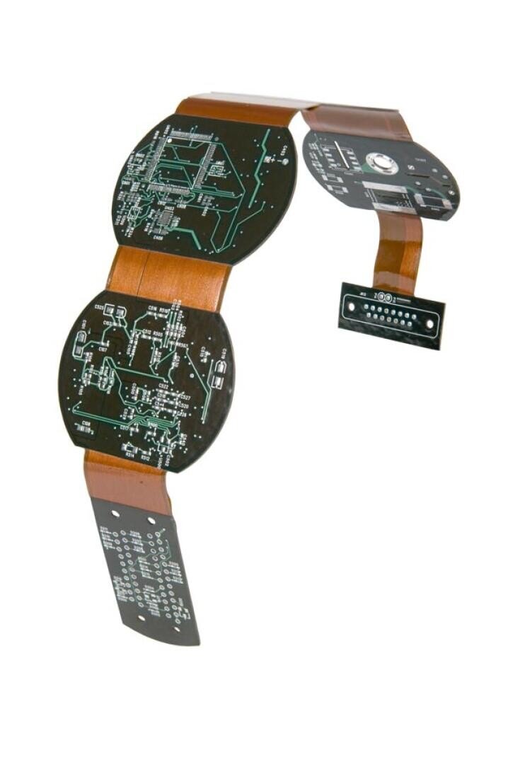 A flexible electronic circuit with circular and rectangular sections, featuring visible white circuitry patterns, bends in an arc shape against a white background.