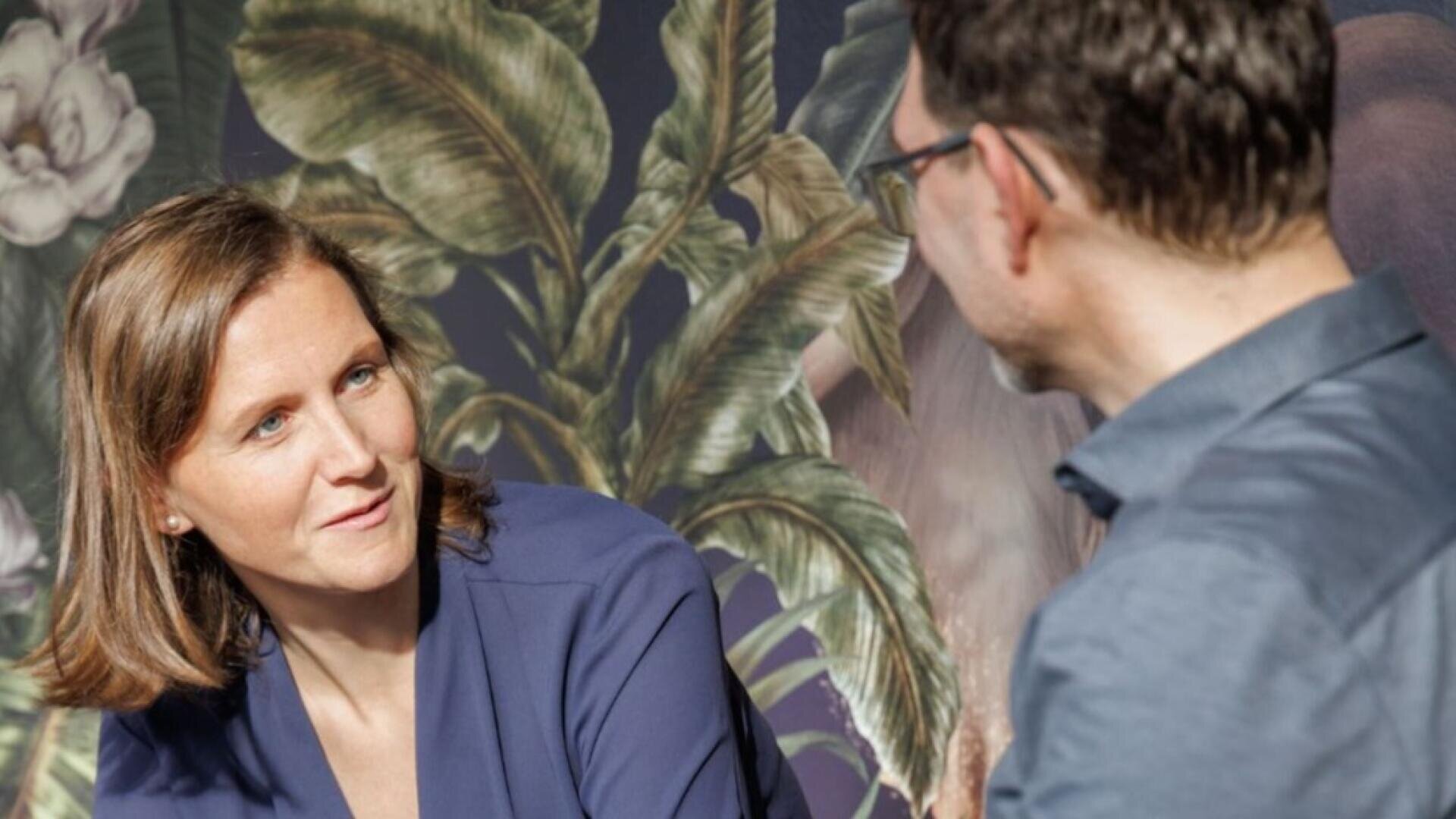 Eine Frau mit schulterlangen braunen Haaren und blauer Bluse hört einem Mann mit Brille und blauem Hemd aufmerksam zu, während im Hintergrund eine Blumentapete zu sehen ist.