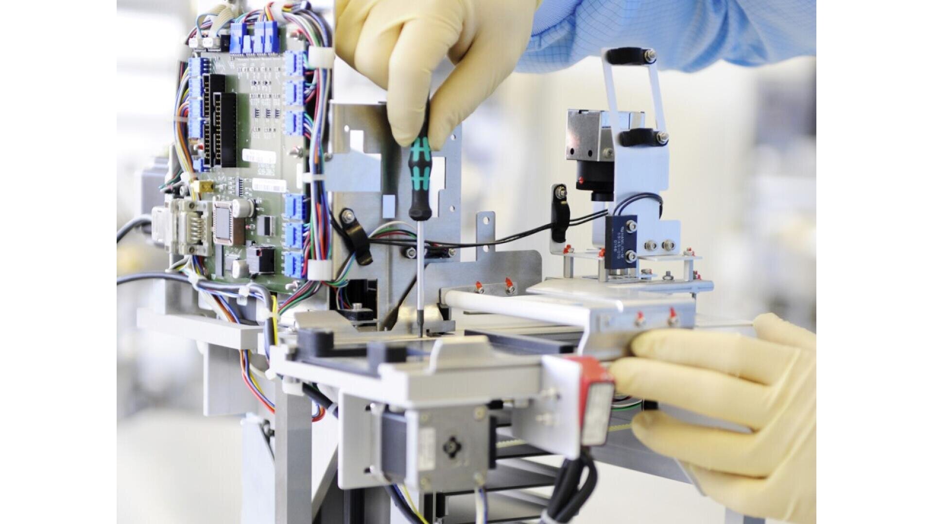 A person wearing latex gloves uses a screwdriver to adjust electronic components in an open, complex machine with circuit boards and wires.