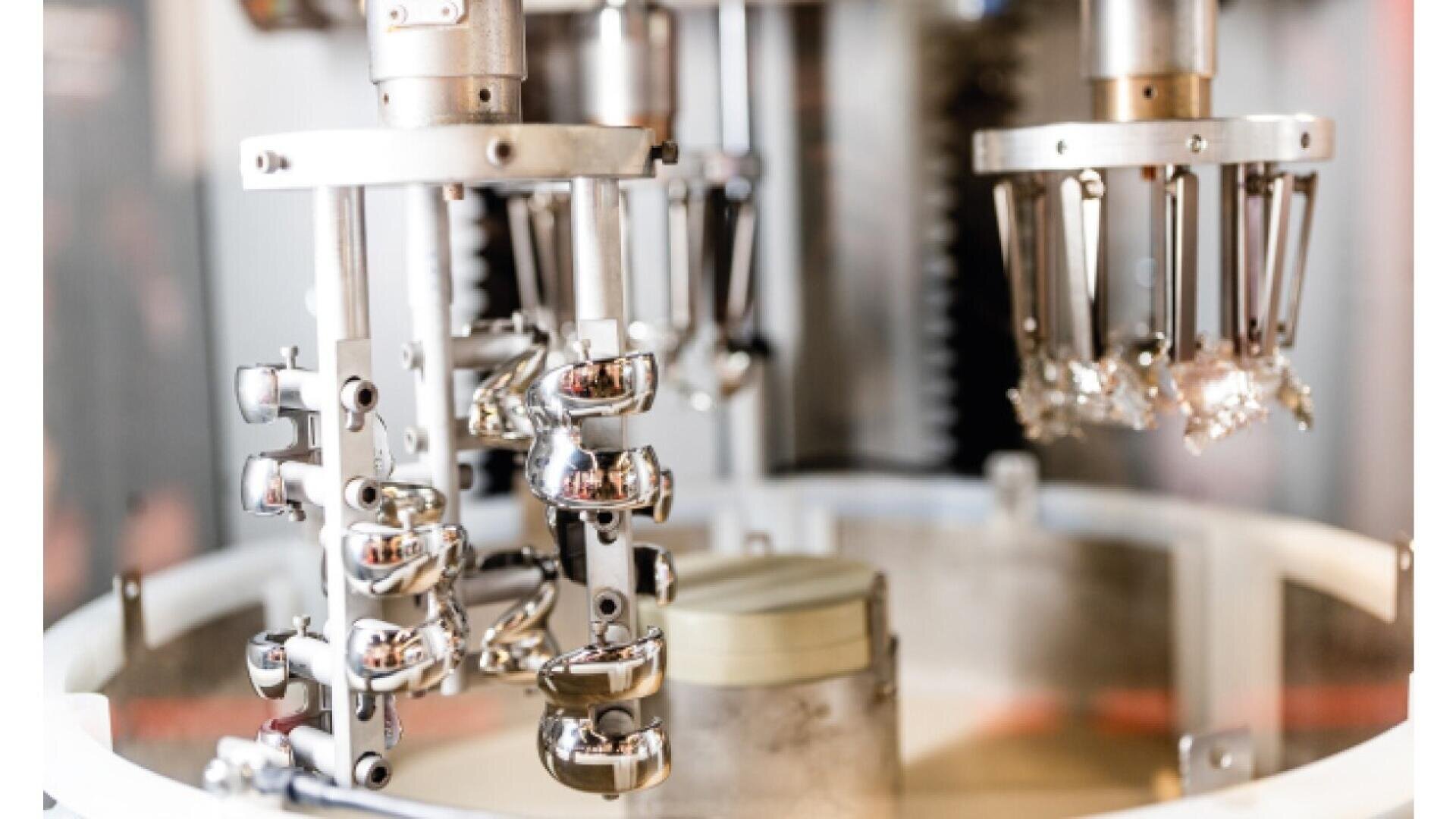 Close-up of a modern industrial machine with shiny metal robotic arms and components, possibly used for automated manufacturing or assembly in a factory setting. The background is blurred.