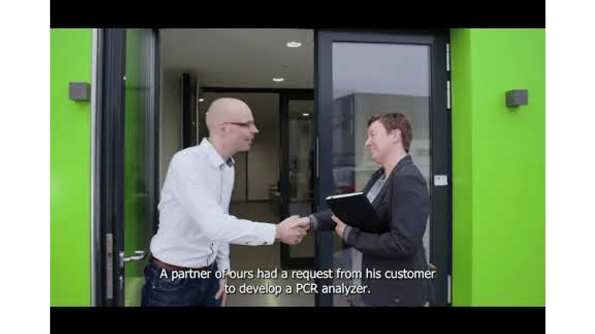 Two people in business attire shake hands outside a building with bright green walls. One holds a folder. The subtitle reads: “A partner of ours had a request from his customer to develop a PCR analyzer.”.