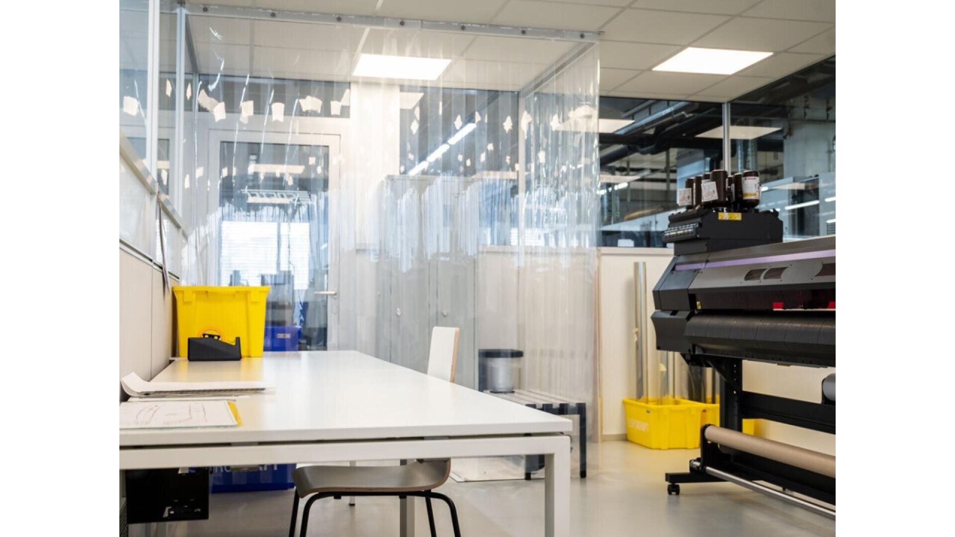 A clean, modern laboratory or workspace features a white table with a chair, a large printer or industrial machine, several yellow bins, and transparent plastic curtains dividing the room.