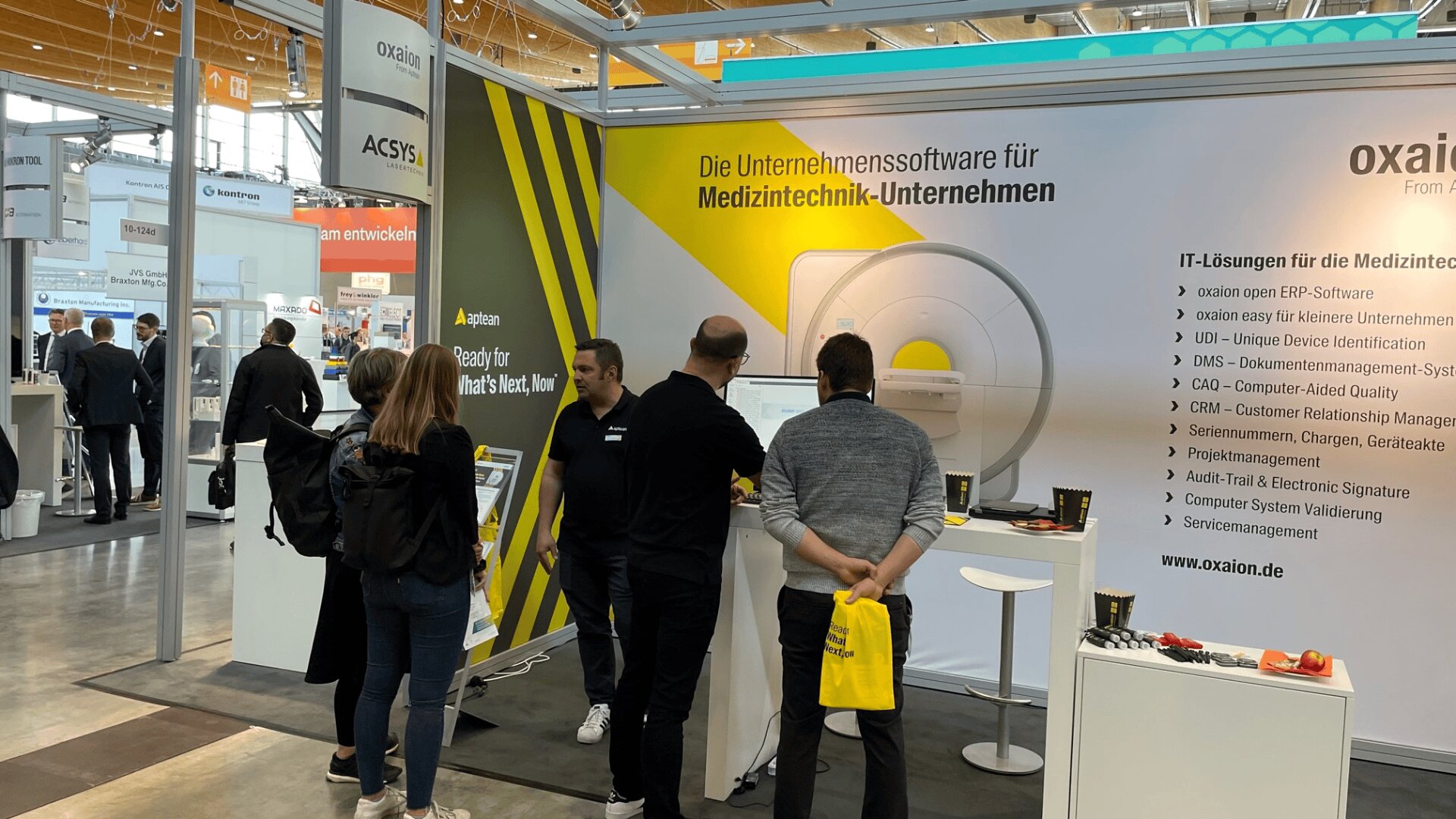 Mehrere Personen stehen am Messestand von oxaiion und unterhalten sich. Ein großes gelb-weißes Banner wirbt für Business-Software für Medizintechnikunternehmen. Ein Display und Werbeartikel sind zu sehen.