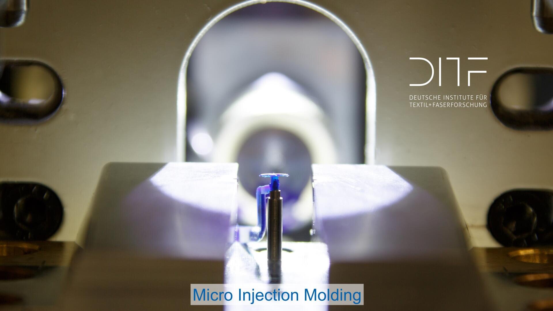 Close-up of a micro injection molding machine with a small blue plastic component in the center, illuminated by bright lights. The DITF logo appears in the top right corner. The caption reads Micro Injection Molding.