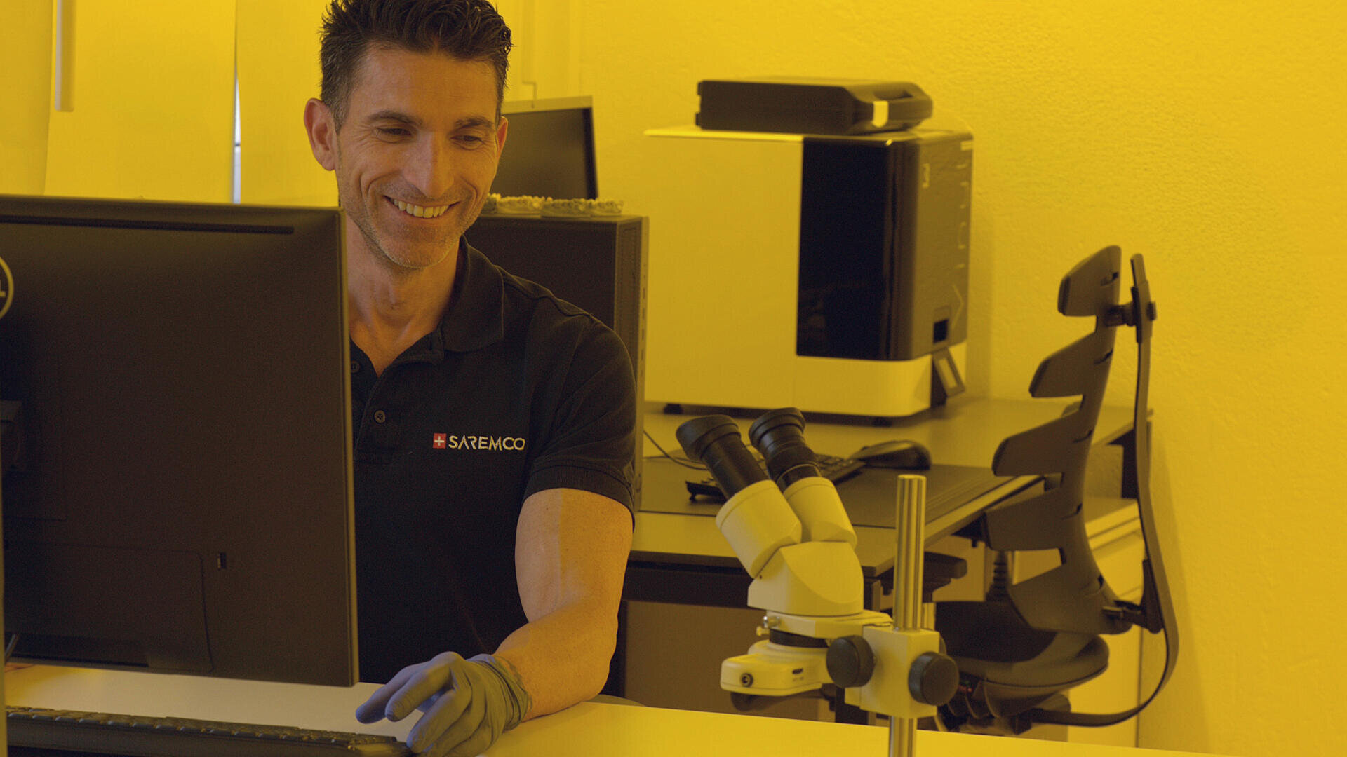 Ein Mann mit Handschuhen und schwarzem Polohemd sitzt an einem Schreibtisch mit Computermonitor und Mikroskop in einem gelb beleuchteten Raum und lächelt bei der Arbeit.