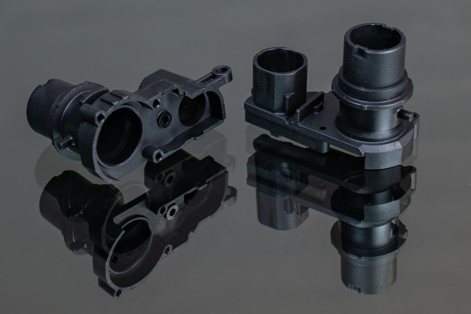 Two black mechanical plastic parts with cylindrical and angular features are placed on a reflective, dark surface so that clear reflections appear under each part.