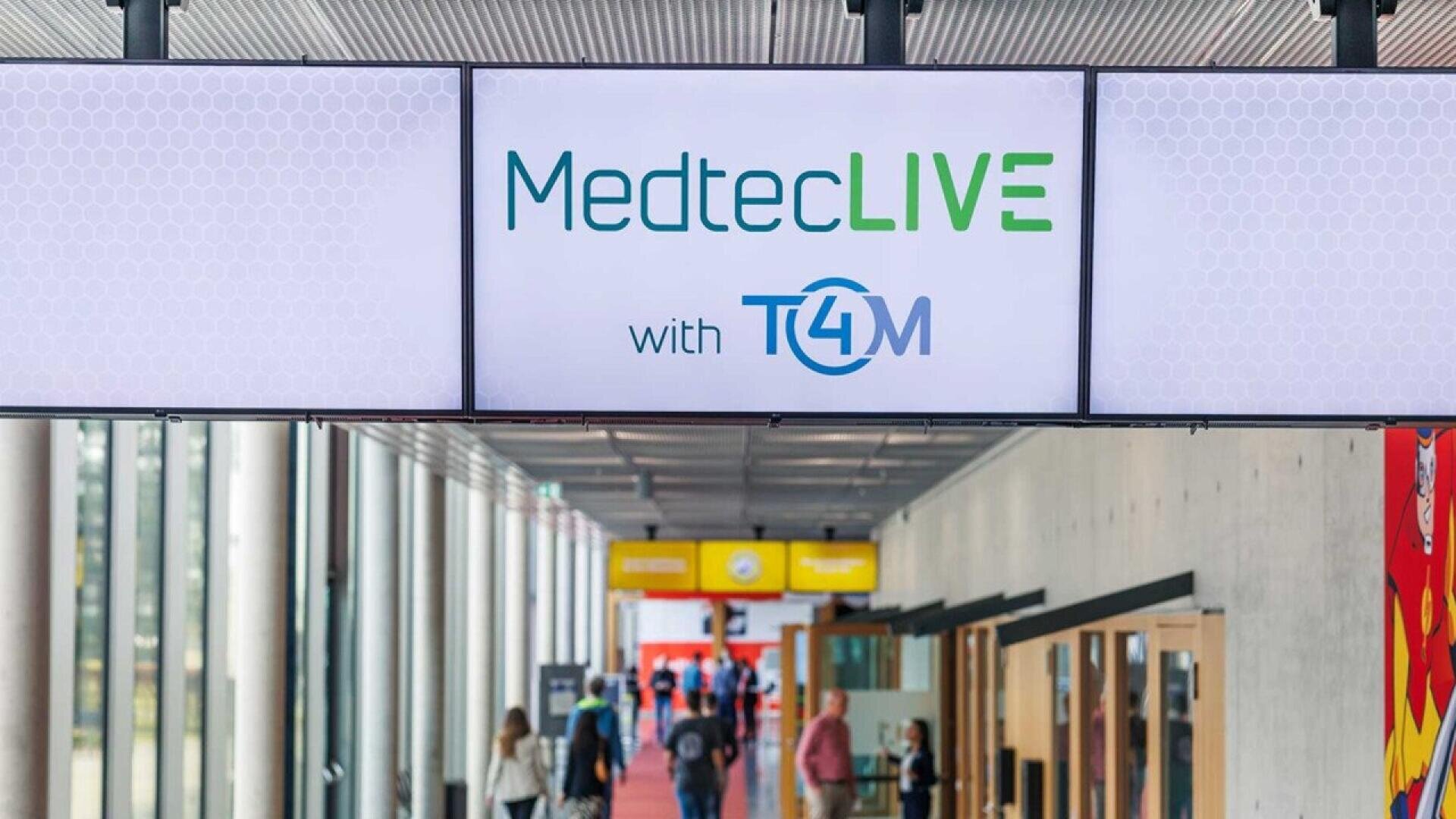 A digital sign displays MedtecLIVE with T4M above a hallway where people are walking, with glass walls and colorful banners visible in the background.
