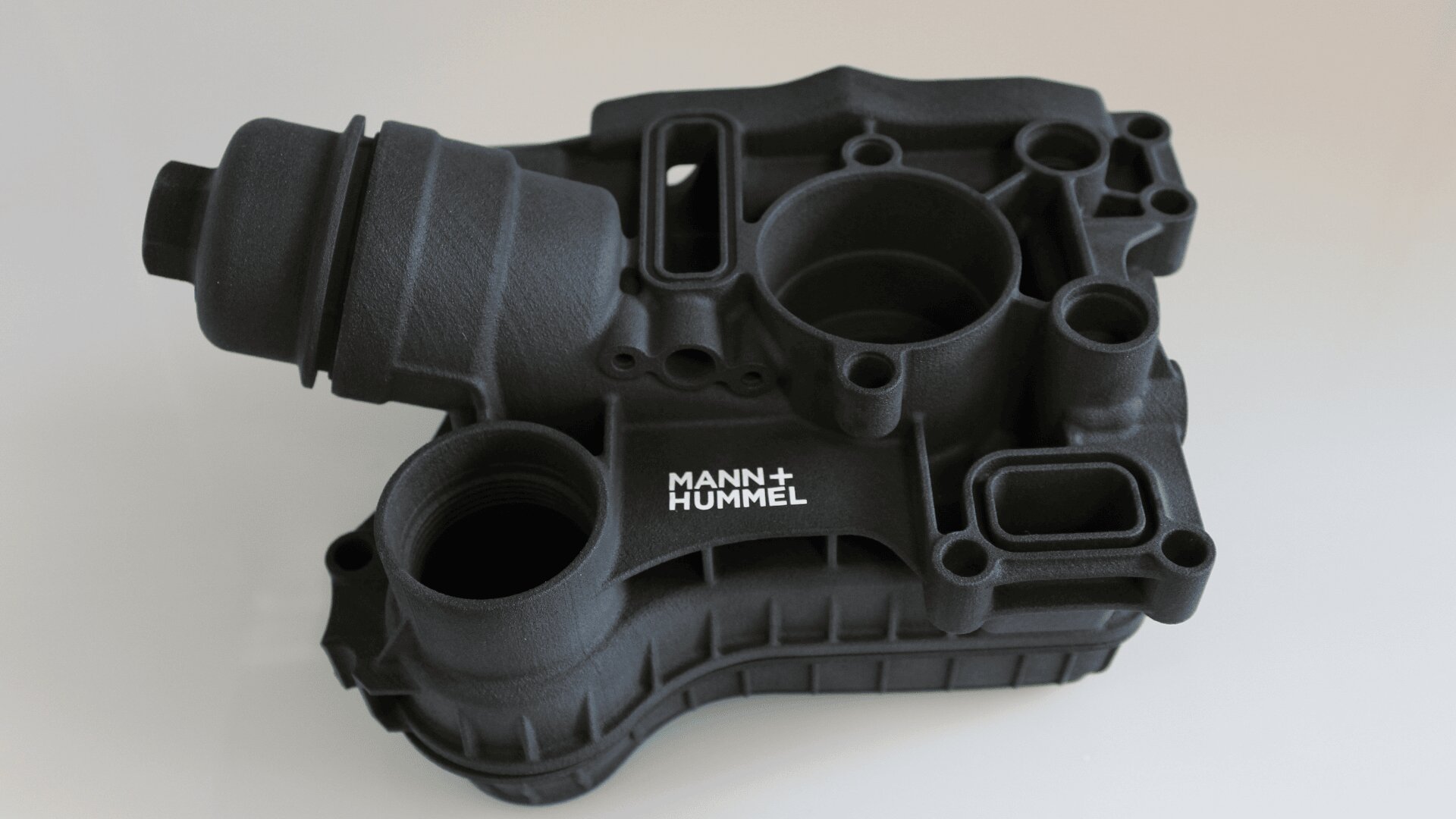 Ein schwarzes 3D-gedrucktes Automobilteil mit zylindrischem und rechteckigem Querschnitt, mit dem weißen MANN+HUMMEL Logo an der Seite, auf einem schlichten hellen Hintergrund.