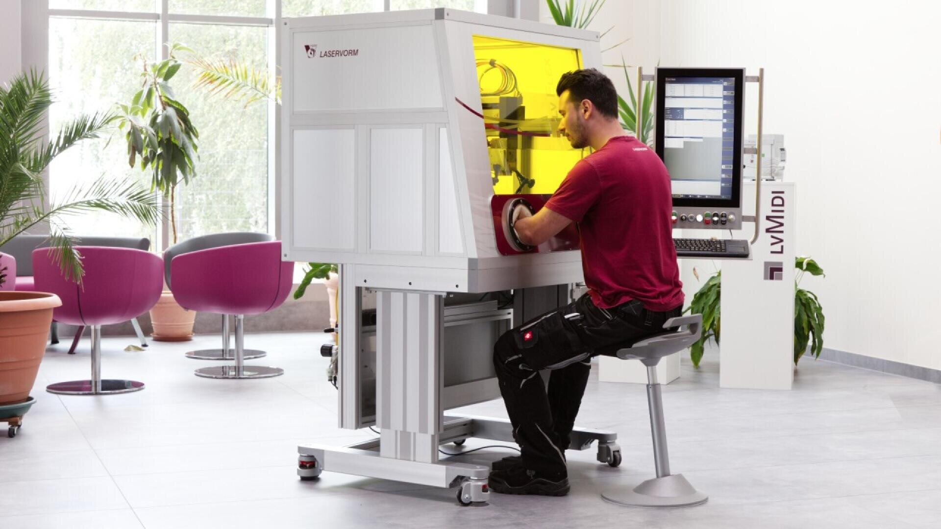 A man sits on a stool operating a white and yellow laser welding machine in a bright, modern room with large windows, plants, and contemporary furniture in the background.