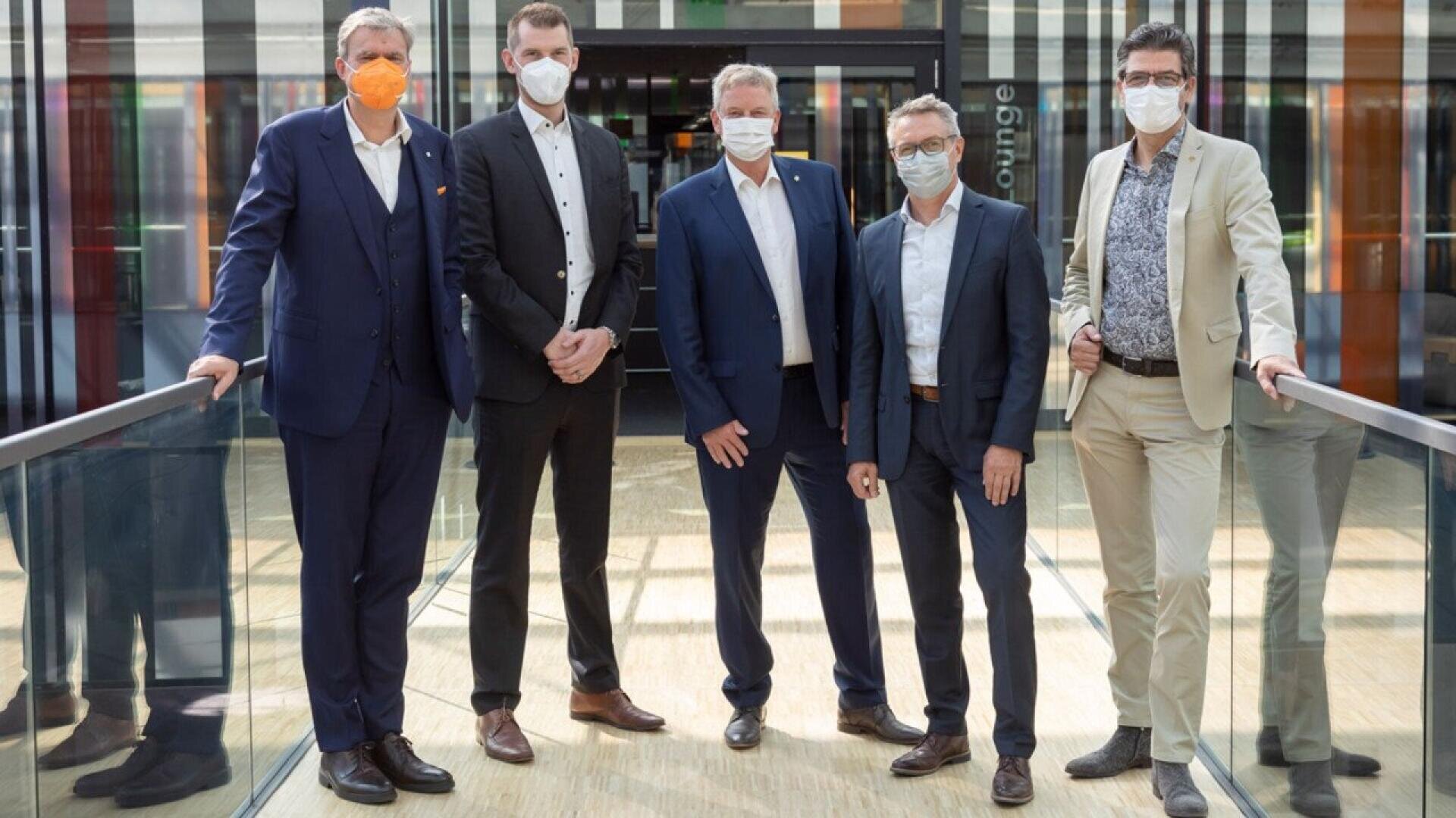 Five men in suits and face masks stand side by side on a glass walkway indoors. They are posing for the photo, with some having their hands in their pockets. The background is modern with glass and colorful panels.