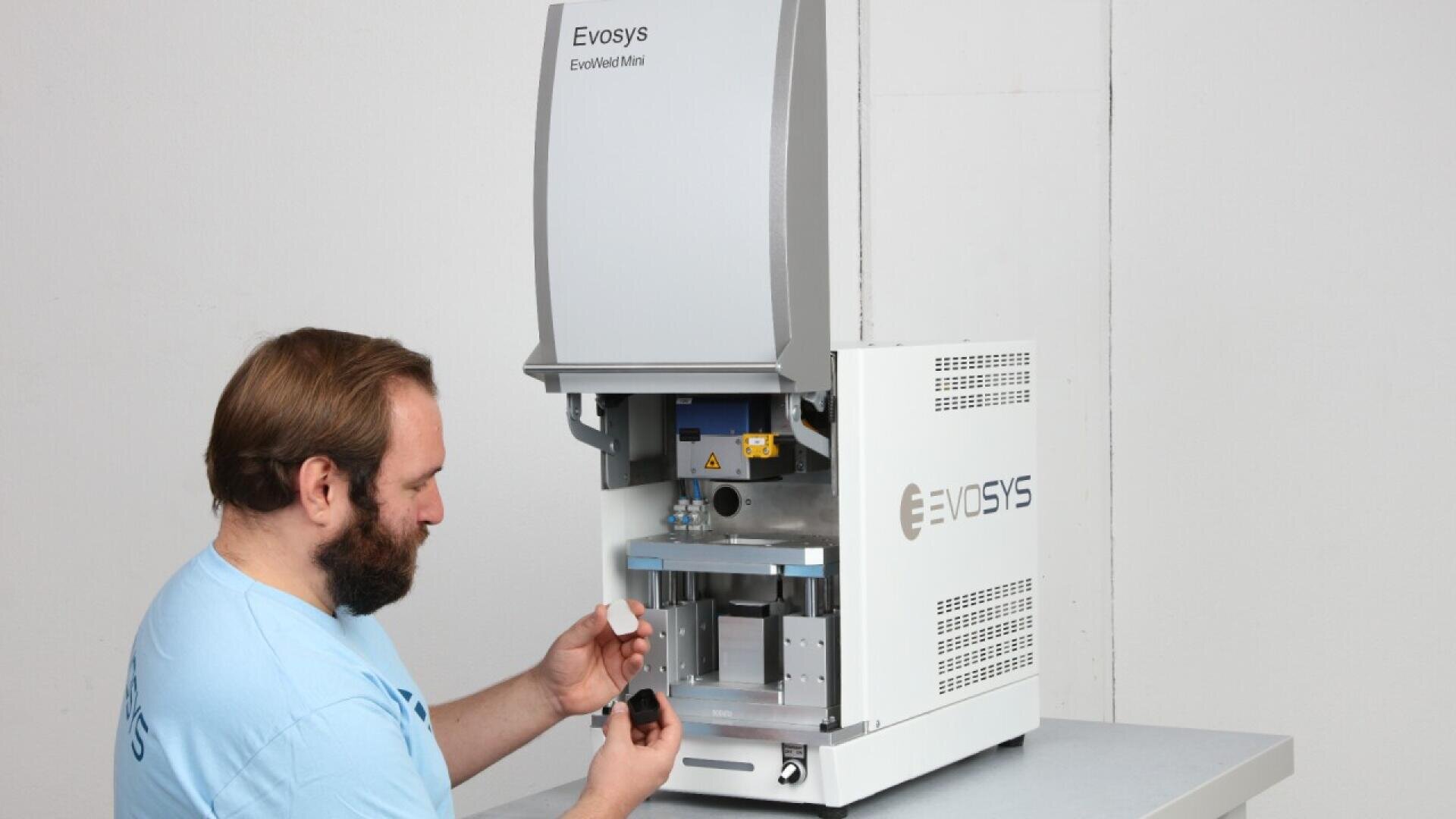 A man in a light blue shirt operates an Evosys EvoWeld Mini machine, examining a small white object in his hand while standing beside the open device on a table.
