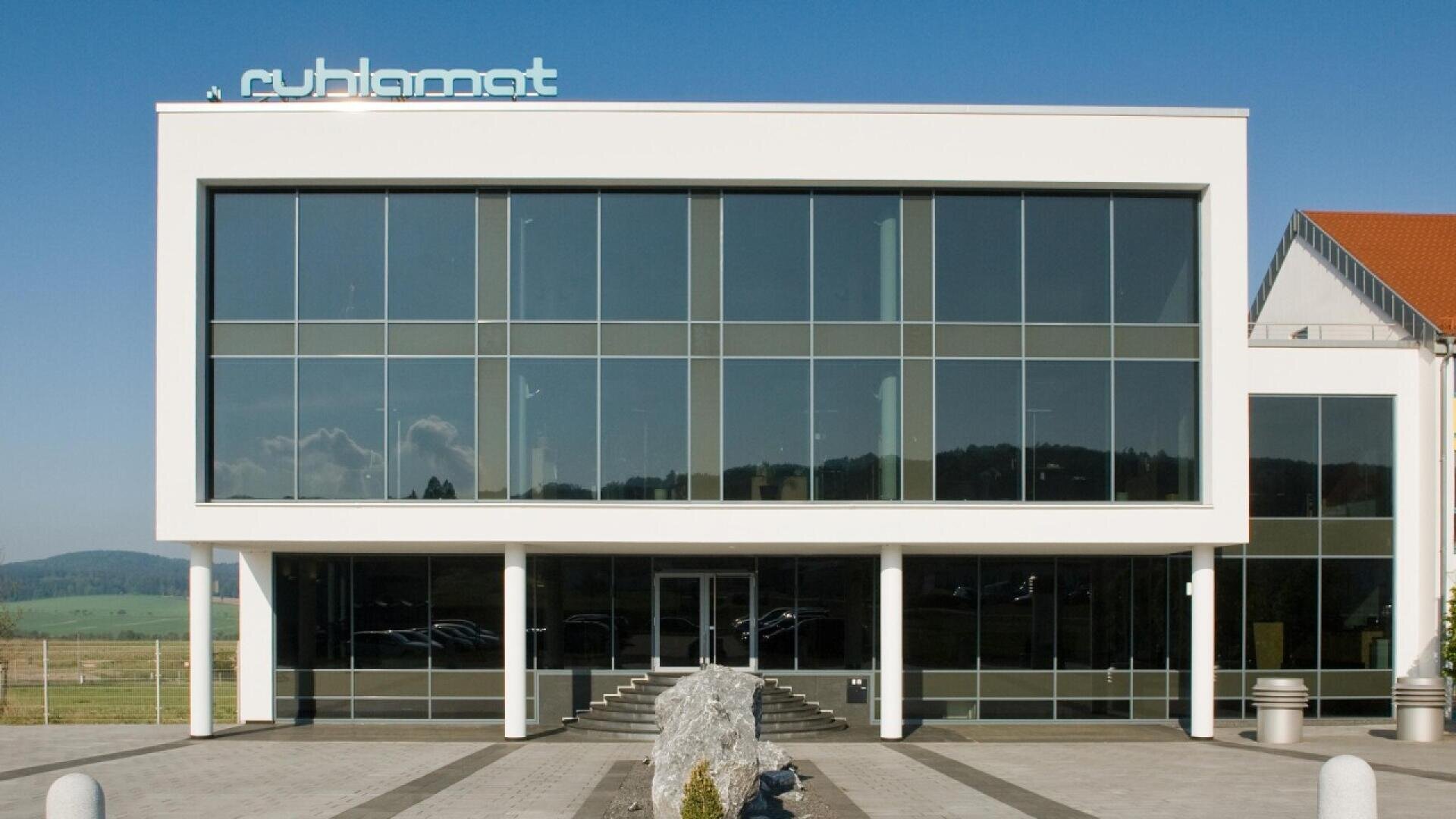 A modern two-story building with large glass windows, a flat roof, and the word cylibomat on top. The entrance has double doors and is flanked by columns, with a landscaped path and rocks in front.