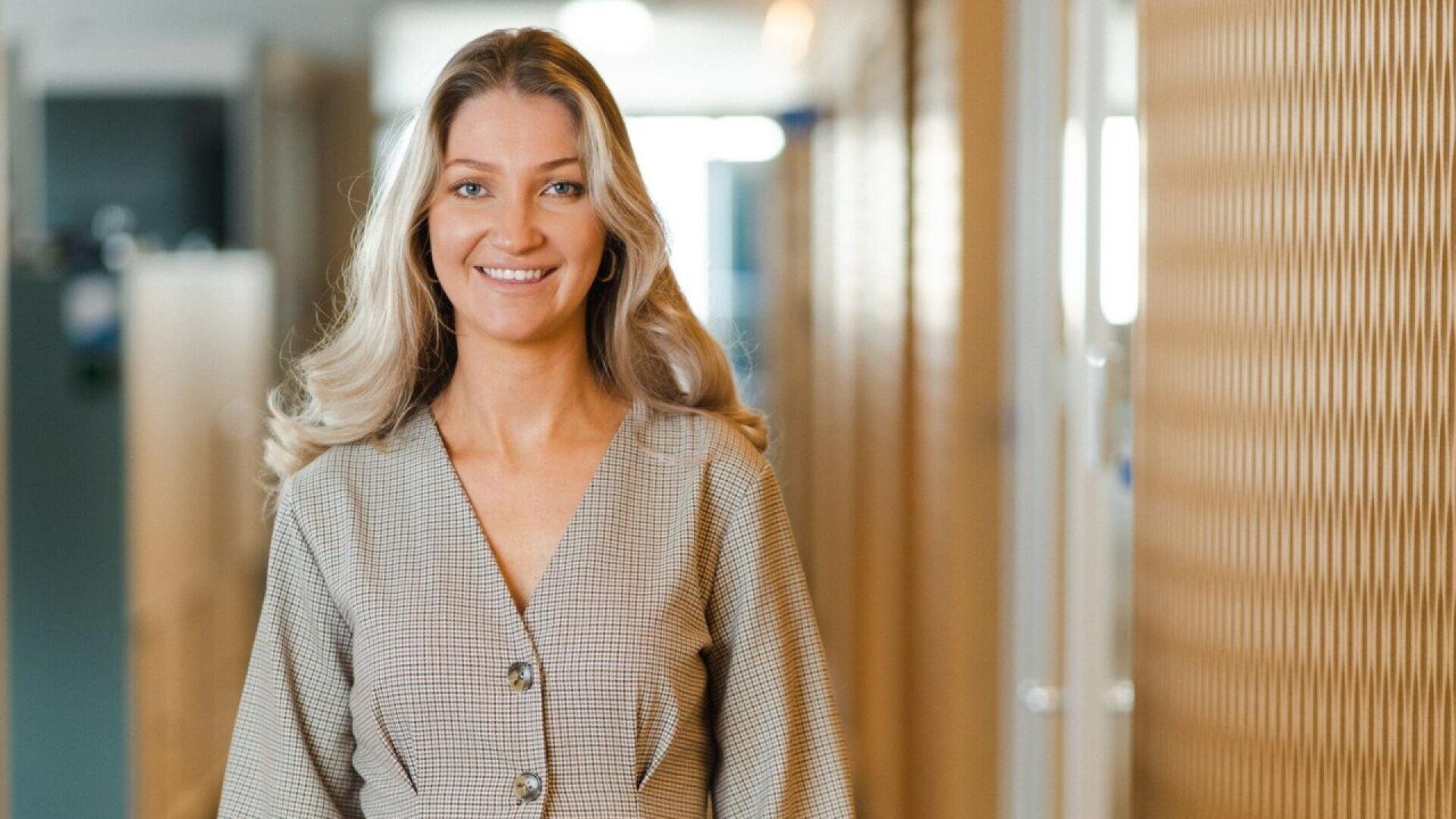 A woman with long, wavy blonde hair smiles at the camera while standing indoors in a hallway with soft lighting and wooden paneling on the right side. She is wearing a beige checkered dress with large buttons.