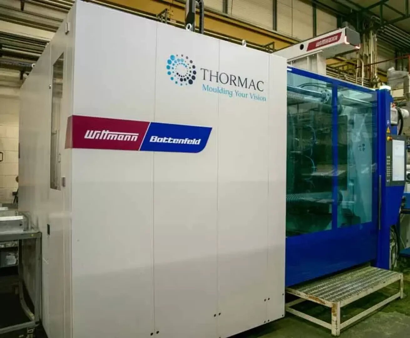 A large industrial injection moulding machine labeled Wittmann Battenfeld and Thormac Moulding Your Vision stands in a factory setting with metal flooring and overhead piping visible.