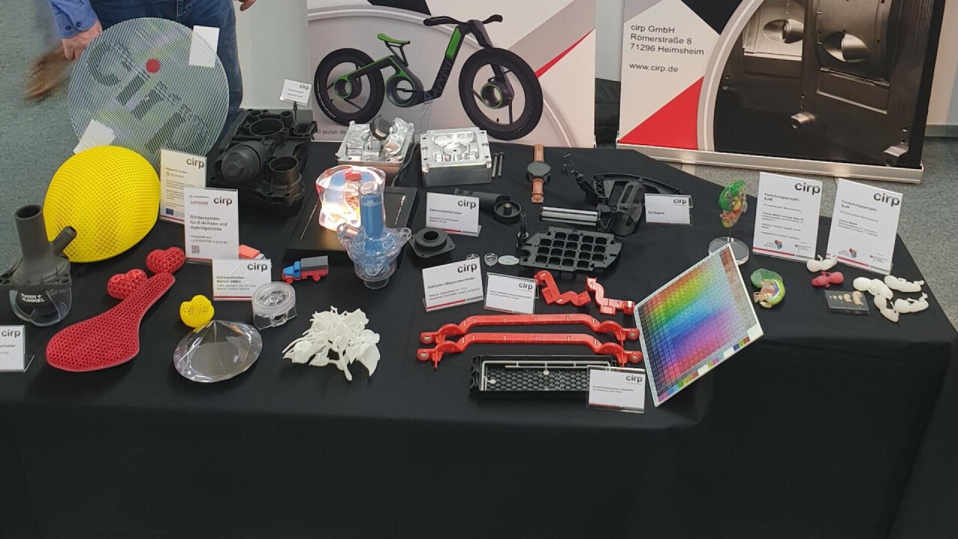 A display table features various 3D-printed objects, models, and parts in different shapes, colors, and sizes, with descriptive cards in front of each item. Banners in the background show a bike and company details.