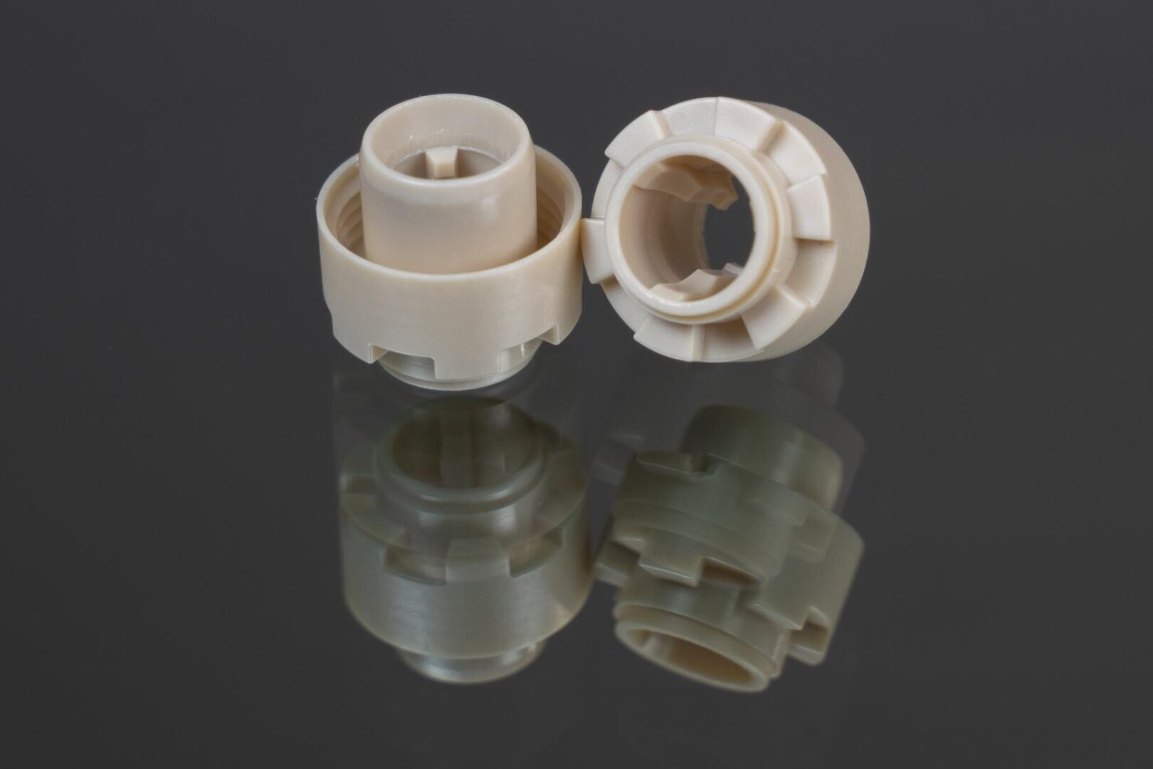Two beige-colored cylindrical plastic parts with inner and outer grooves are depicted on a glossy black surface so that the parts underneath are clearly visible.