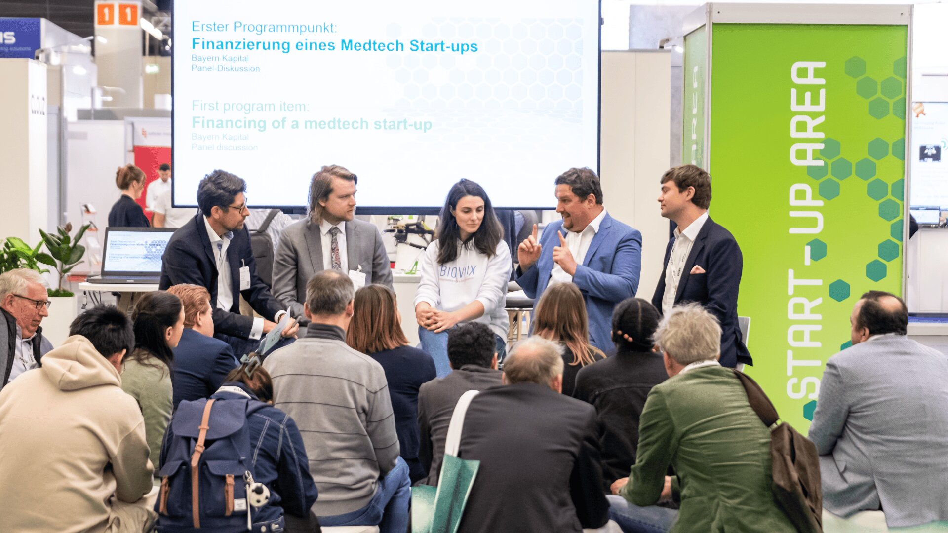 A group of people watch a panel discussion about medtech start-up financing at a conference, with five speakers seated on a stage and an audience sitting closely in front of them. A START-UP AREA sign is visible.