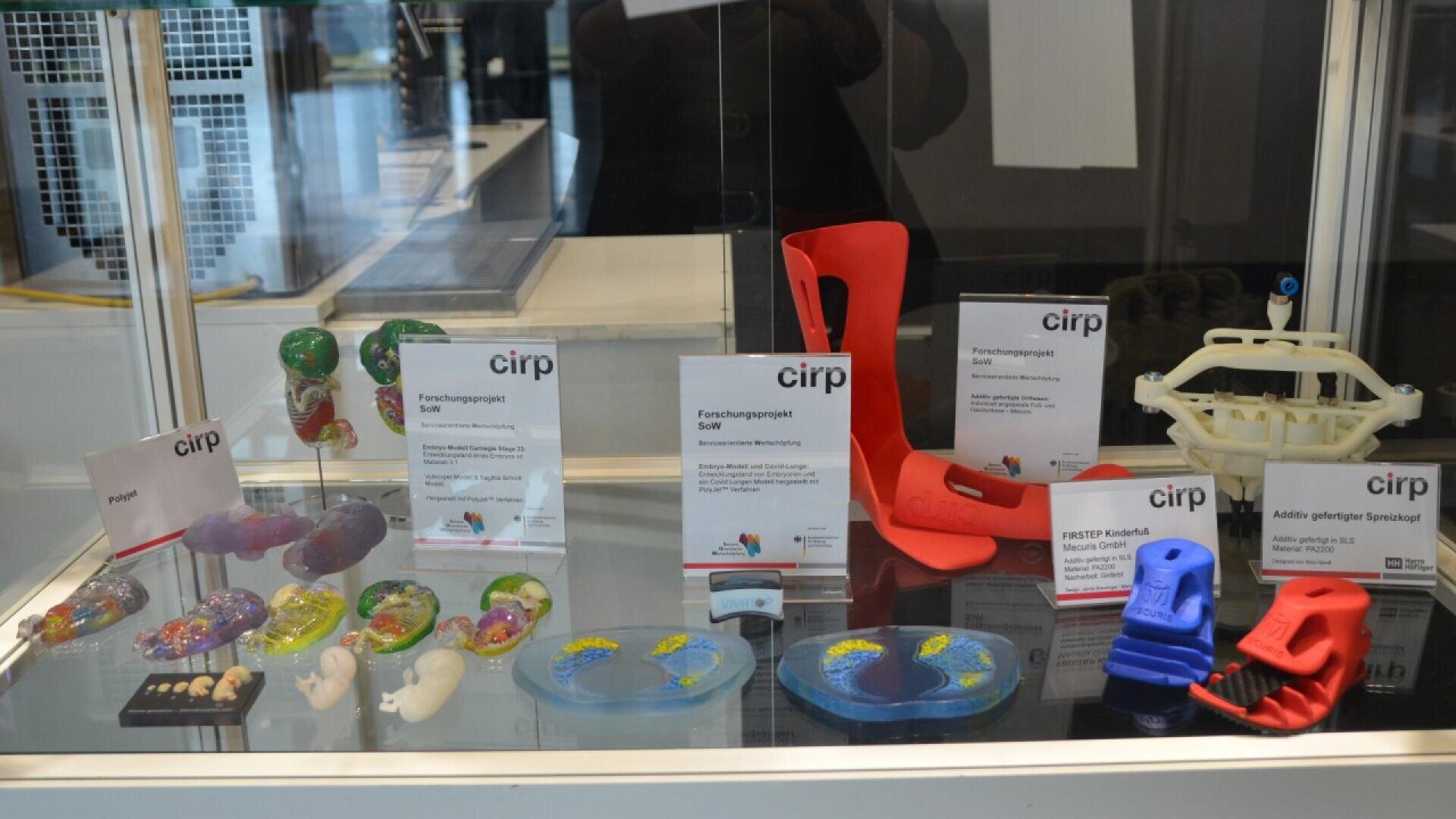 A display case showing various colorful 3D-printed models, anatomical replicas, machine parts, and informational cards labeled “cirp,” representing research projects and prototypes.