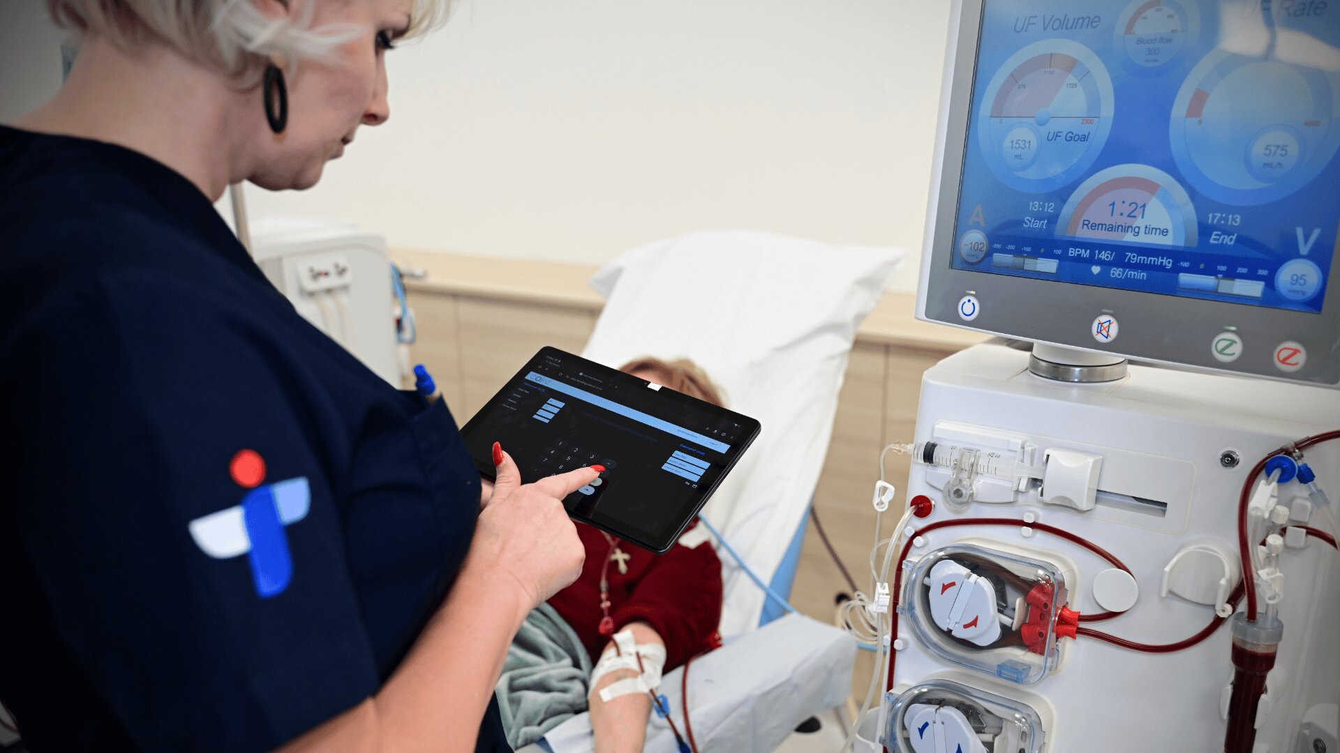 A medical professional uses a tablet to monitor a dialysis machine next to a patient lying in a hospital bed. The machine displays various medical data on its screen.