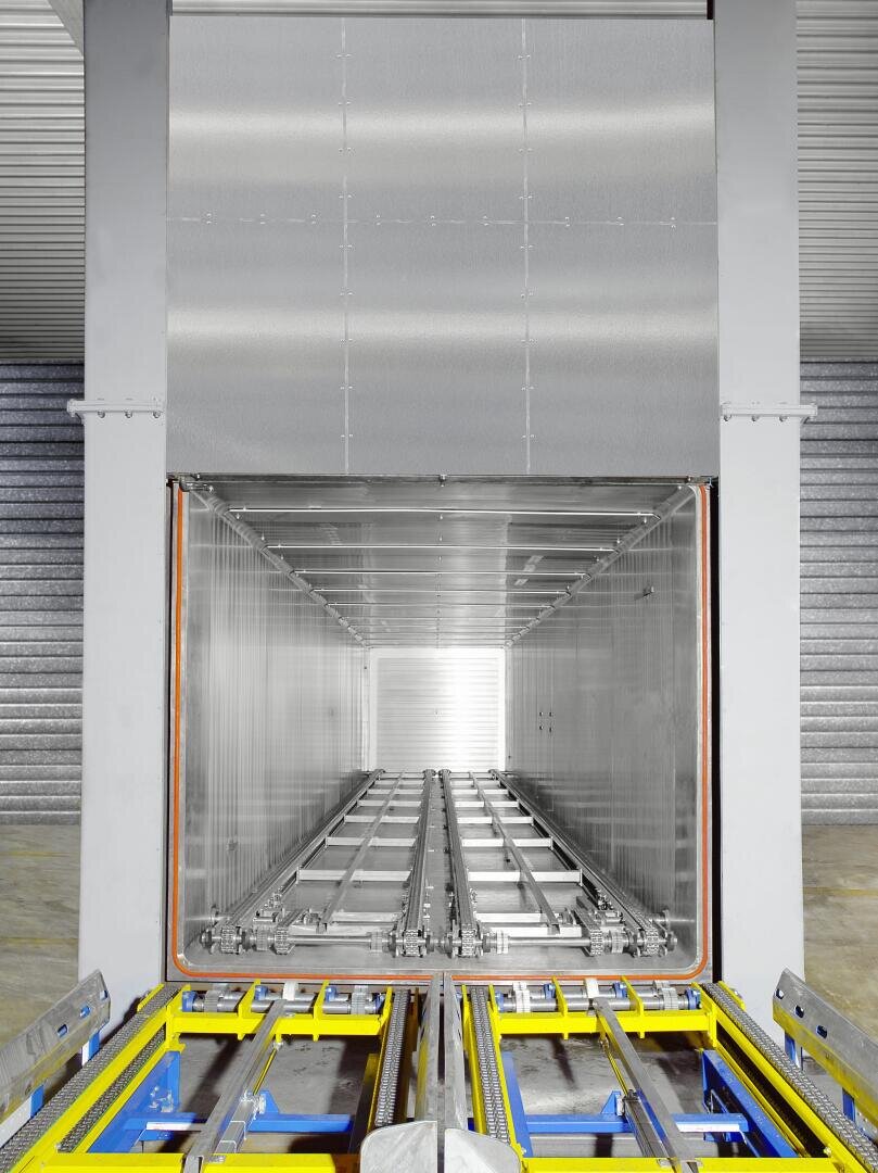 Ein Industrietunnel aus Edelstahl mit Metallschienen und gelben Sicherheitsbarrieren, wahrscheinlich Teil eines Förderbands oder eines automatisierten Ladesystems in einem Lager oder einer Fabrik.