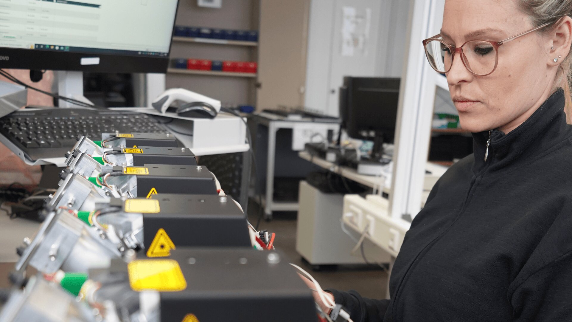 Eine Frau mit Brille und dunkler Jacke prüft elektronische Geräte mit Warnhinweisen auf einer Werkbank in einem Büro oder Labor, wobei neben ihr ein Computermonitor zu sehen ist.