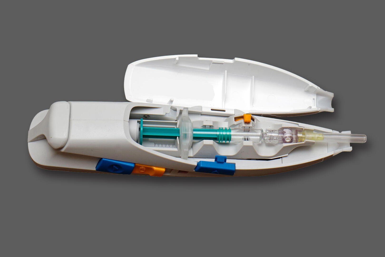 A close-up of an opened medical autoinjector showing its internal components, including a syringe, a needle, and various colored buttons, on a solid gray background.