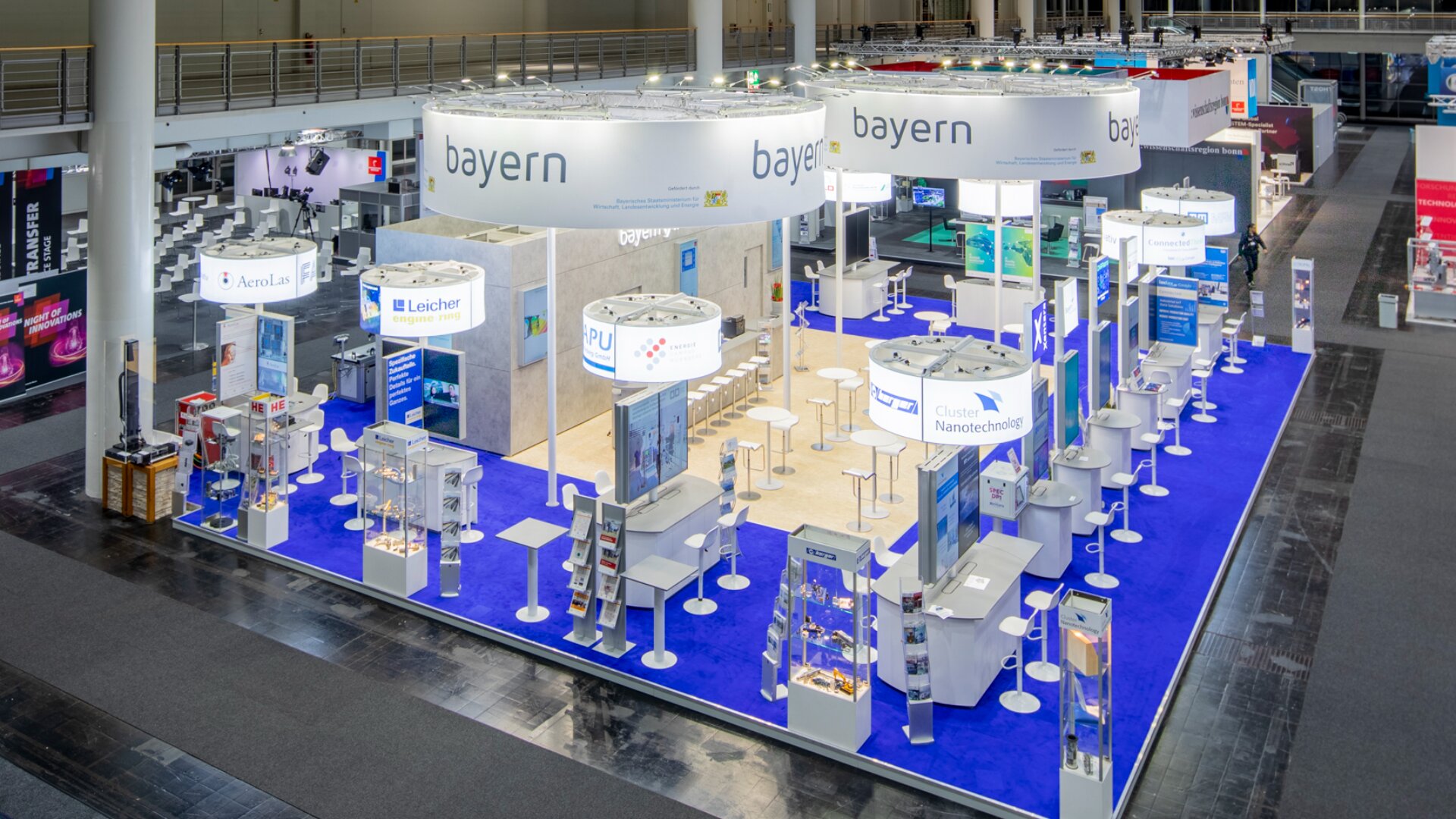 Großer Messestand mit Bayern-Schildern, blauem Teppich, Ausstellungsständen, Broschüren, Sitzgelegenheiten und verschiedenen Technologie- und Wissenschaftspräsentationen in einem geräumigen, gut beleuchteten Kongresszentrum.
