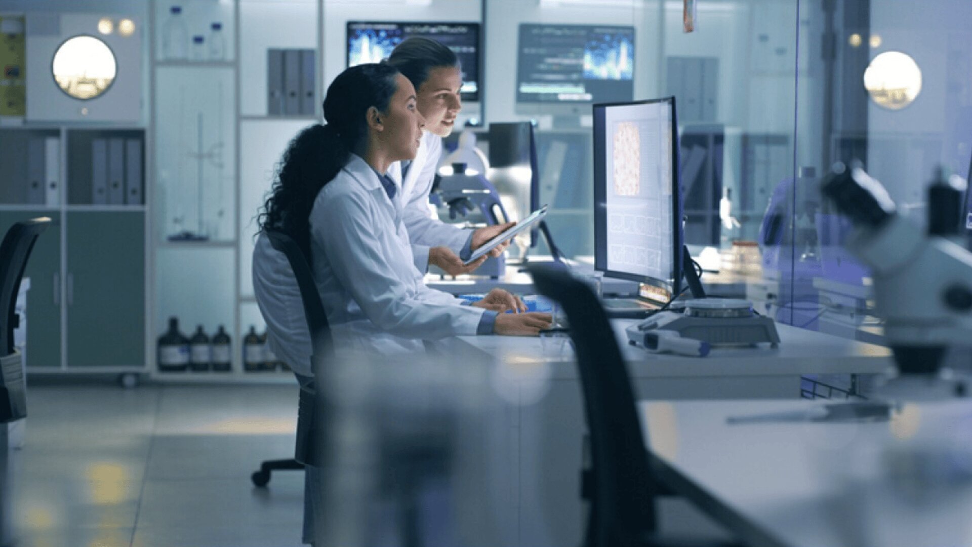 Two scientists in lab coats work together at a computer in a modern laboratory, looking at data on a large screen. Lab equipment and microscopes are visible on the desks around them.