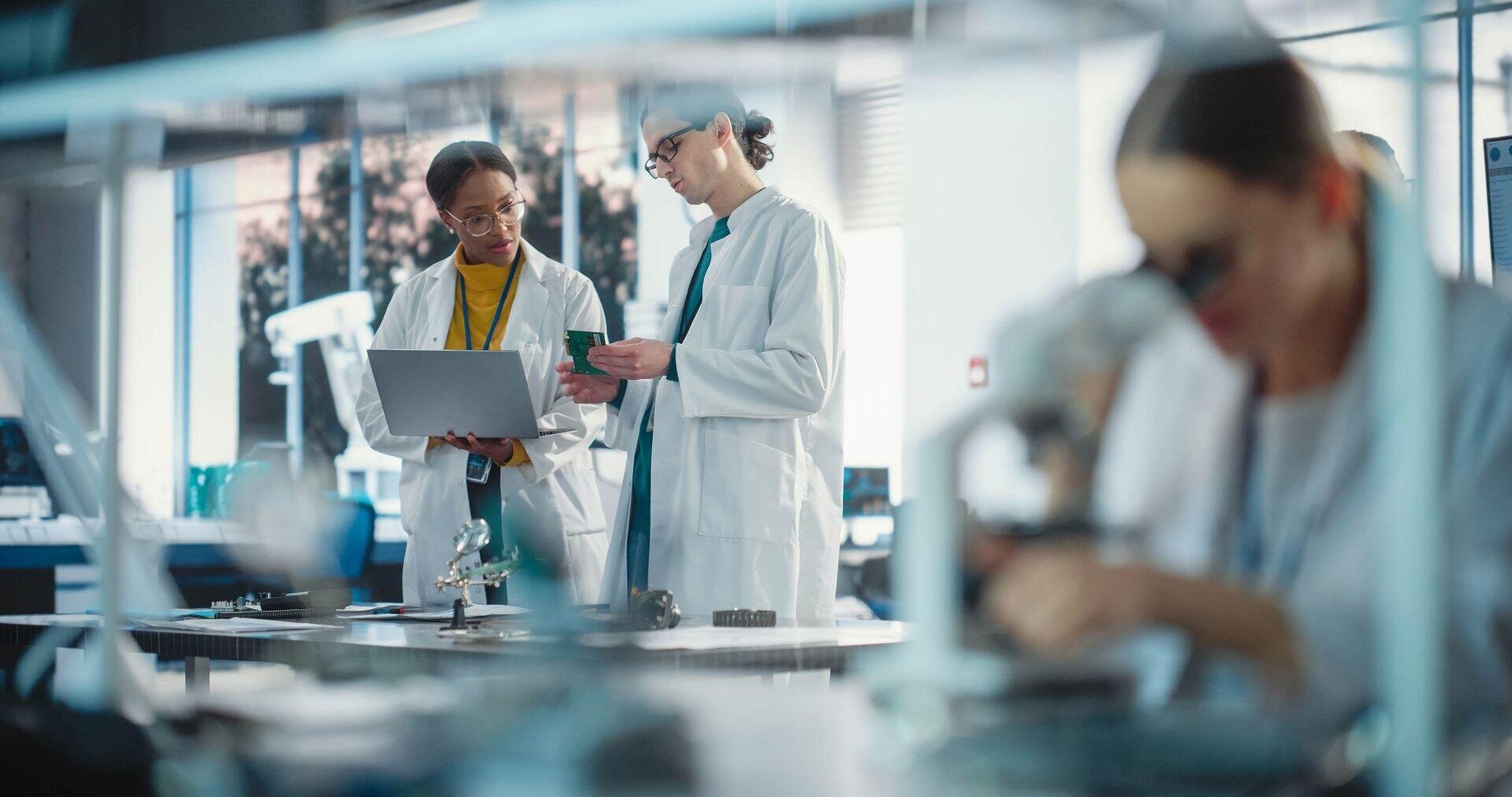 Two scientists in lab coats discuss data on a laptop, while another person in the foreground uses a microscope. Laboratory equipment is visible on tables, creating a busy, modern research environment.