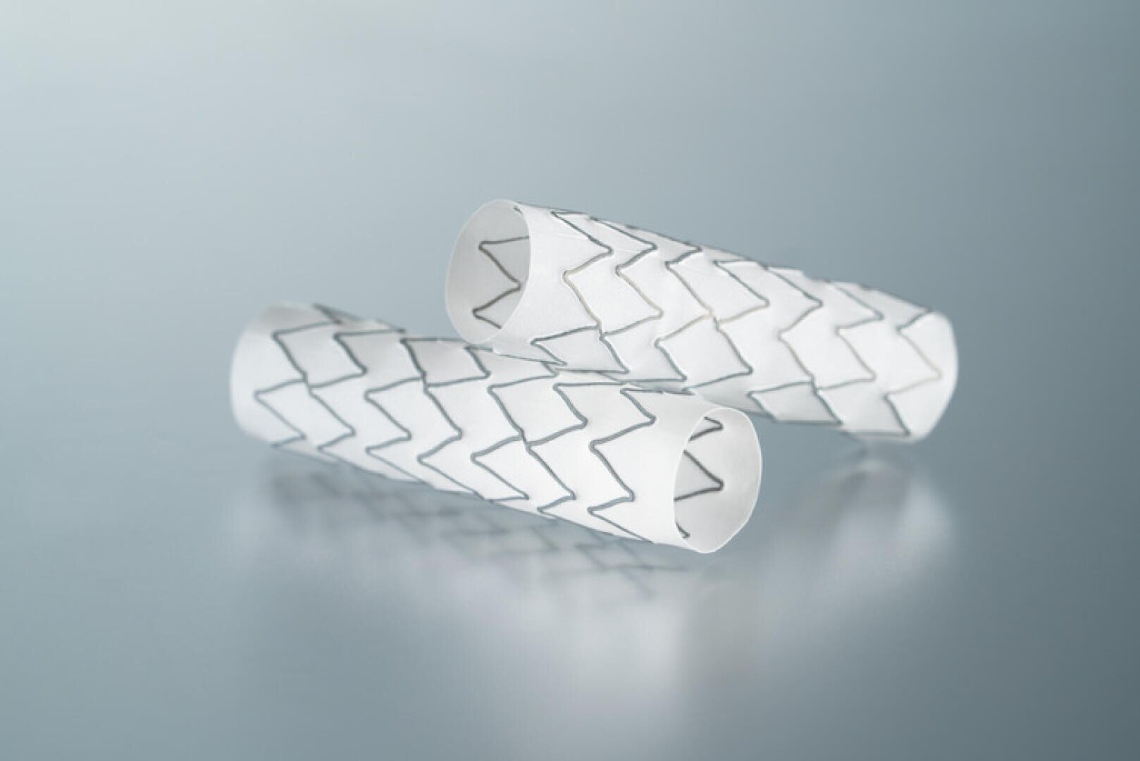 Zwei zylindrische medizinische Stents mit weißer Oberfläche und zickzackförmigen schwarzen Linien sind auf einer glatten, hellblauen Oberfläche platziert.