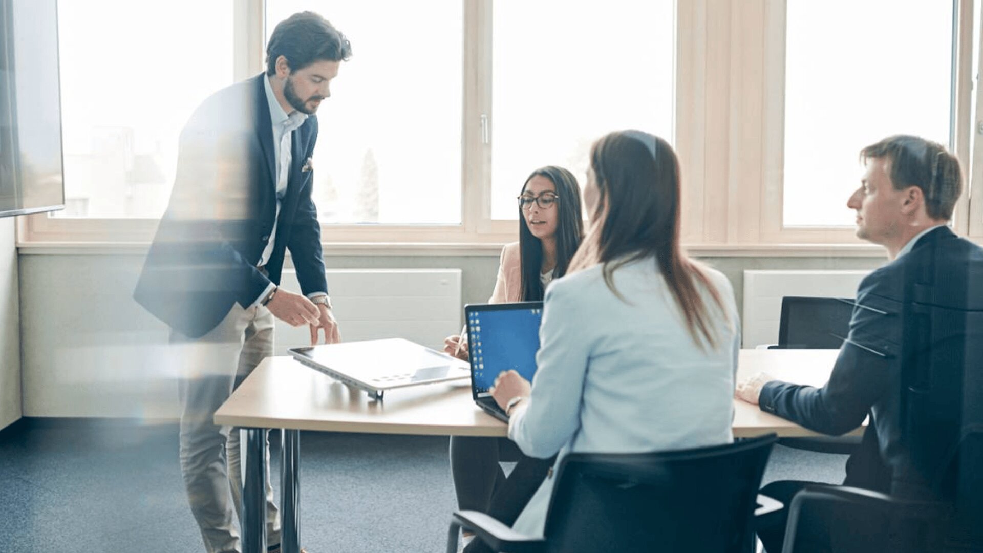 Vier Personen in Geschäftskleidung haben eine Besprechung in einem hellen Büro. Ein Mann steht und deutet auf einen Laptop, während die anderen um einen Tisch mit Laptops und Dokumenten sitzen und zuhören.