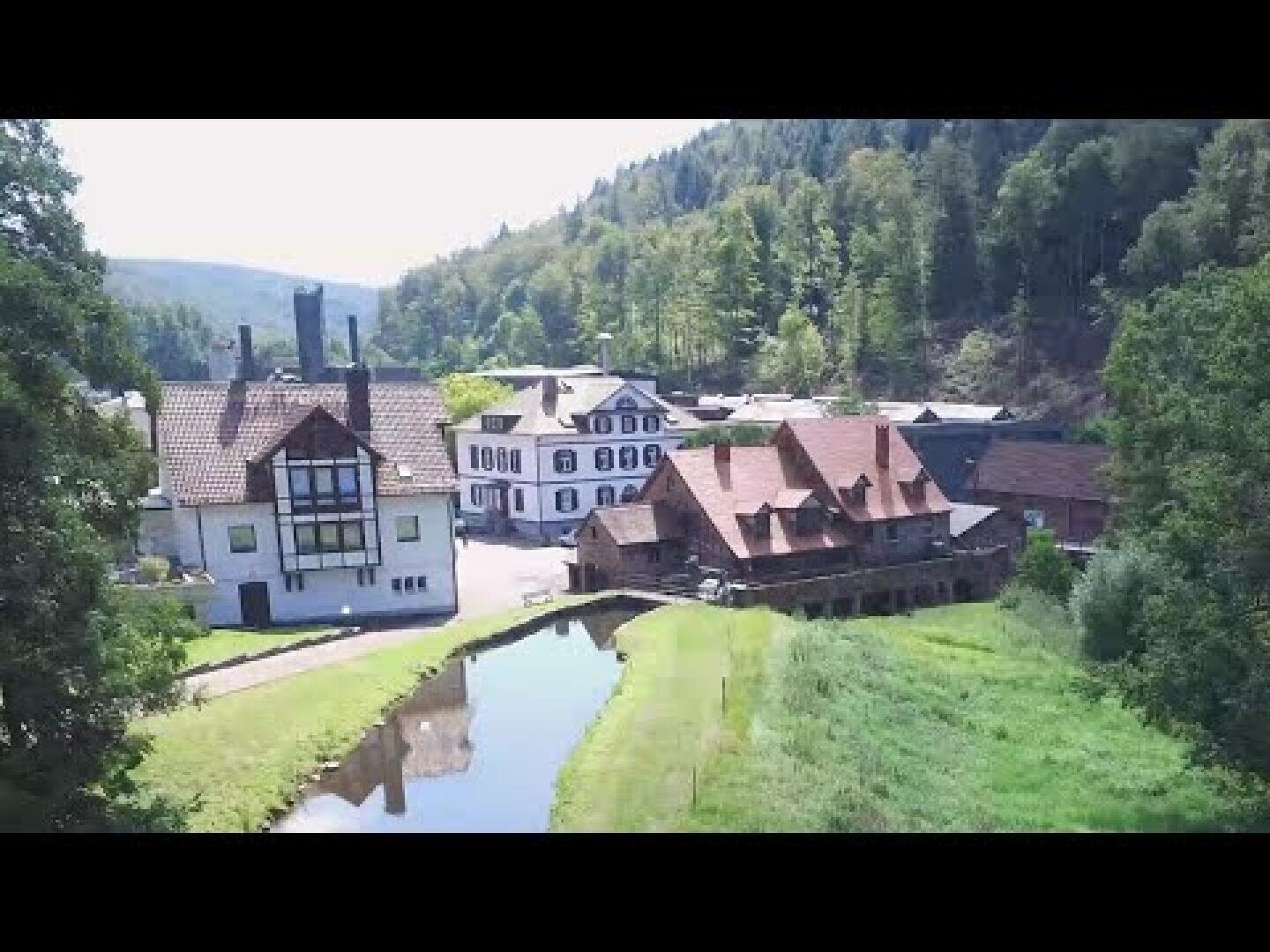 Eine Ansammlung von Gebäuden im traditionellen europäischen Stil mit schrägen Dächern liegt an einem schmalen Fluss, umgeben von grünem Gras, Bäumen und bewaldeten Hügeln an einem sonnigen Tag.