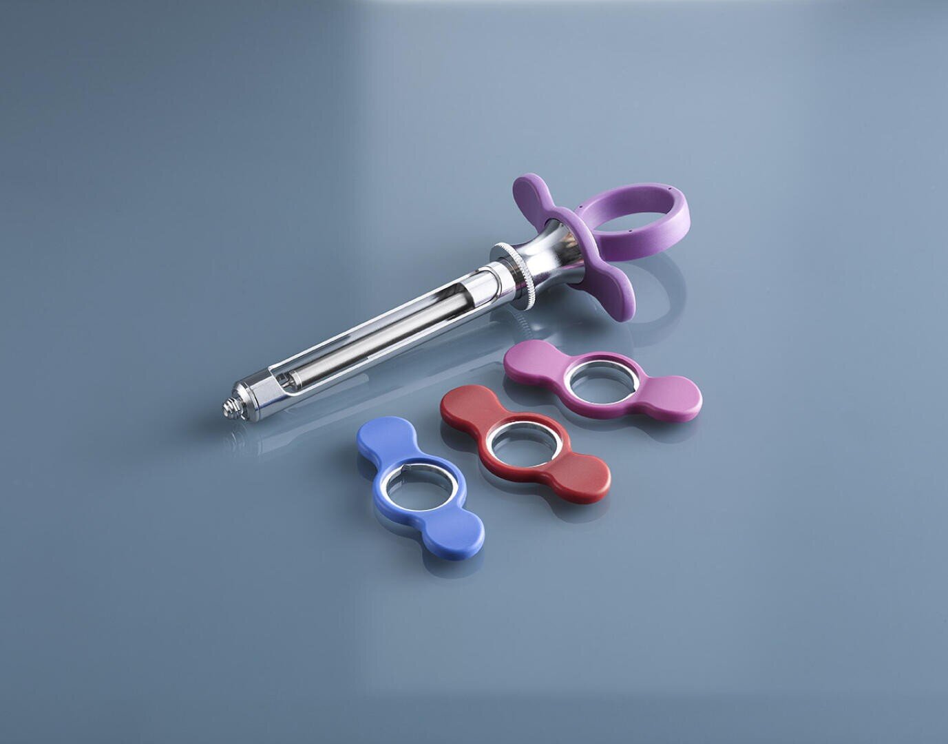A silver dental syringe with a purple butterfly-shaped handle is displayed on a glossy blue surface, alongside three additional butterfly-shaped handles in blue, red, and pink.