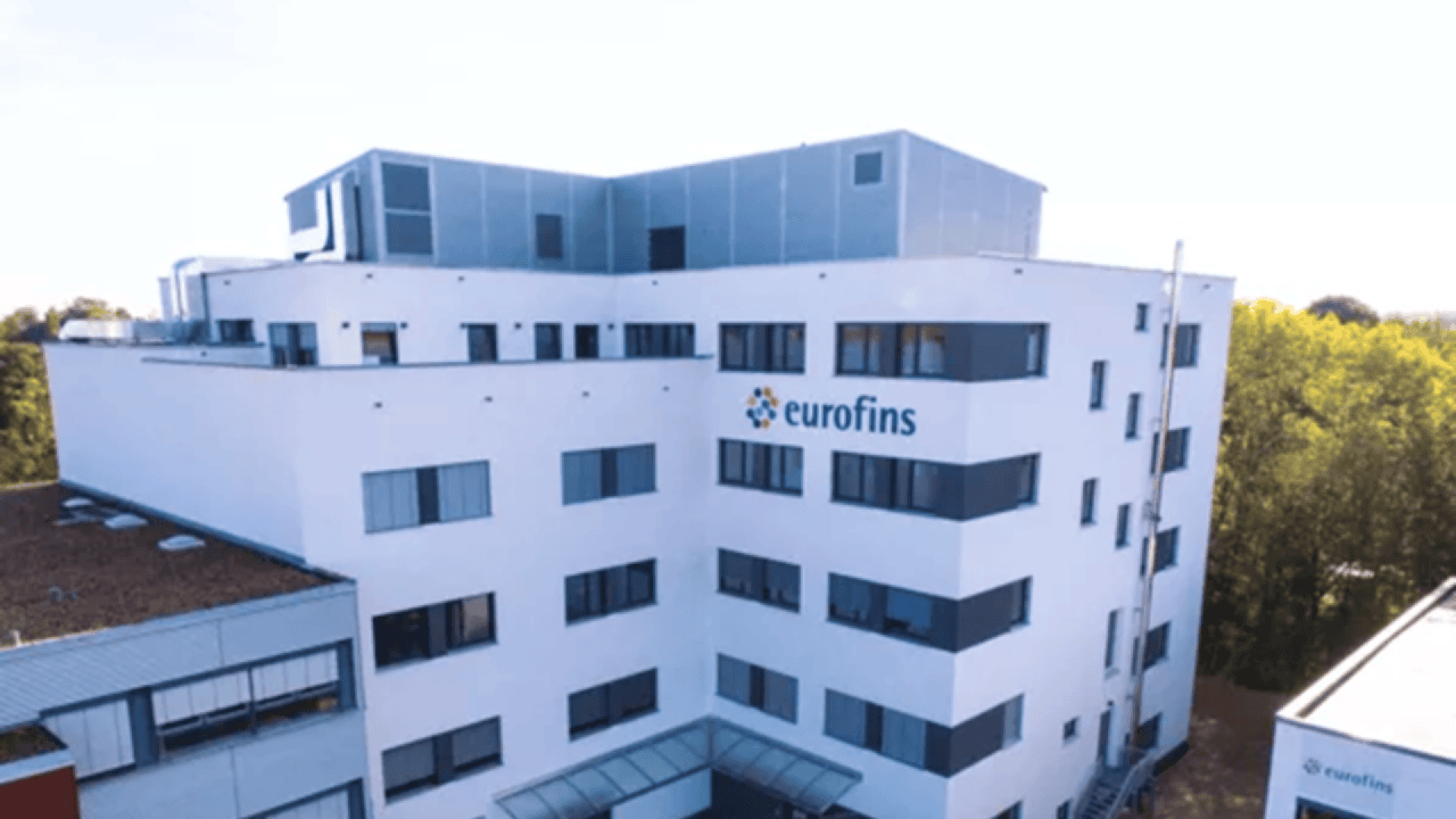 A modern white multi-story building with large windows, displaying the eurofins logo on its facade, surrounded by trees and a clear sky in the background.