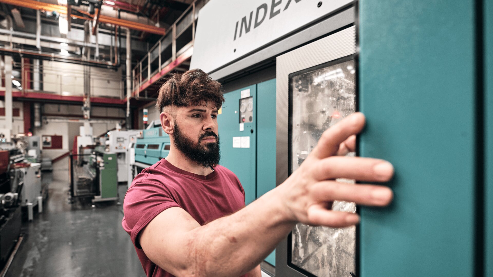 Ein Mann mit Bart und braunem Haar, der ein rotes T-Shirt trägt, bedient eine Schalttafel an einer Industrieanlage in einer großen, modernen Fabrik.
