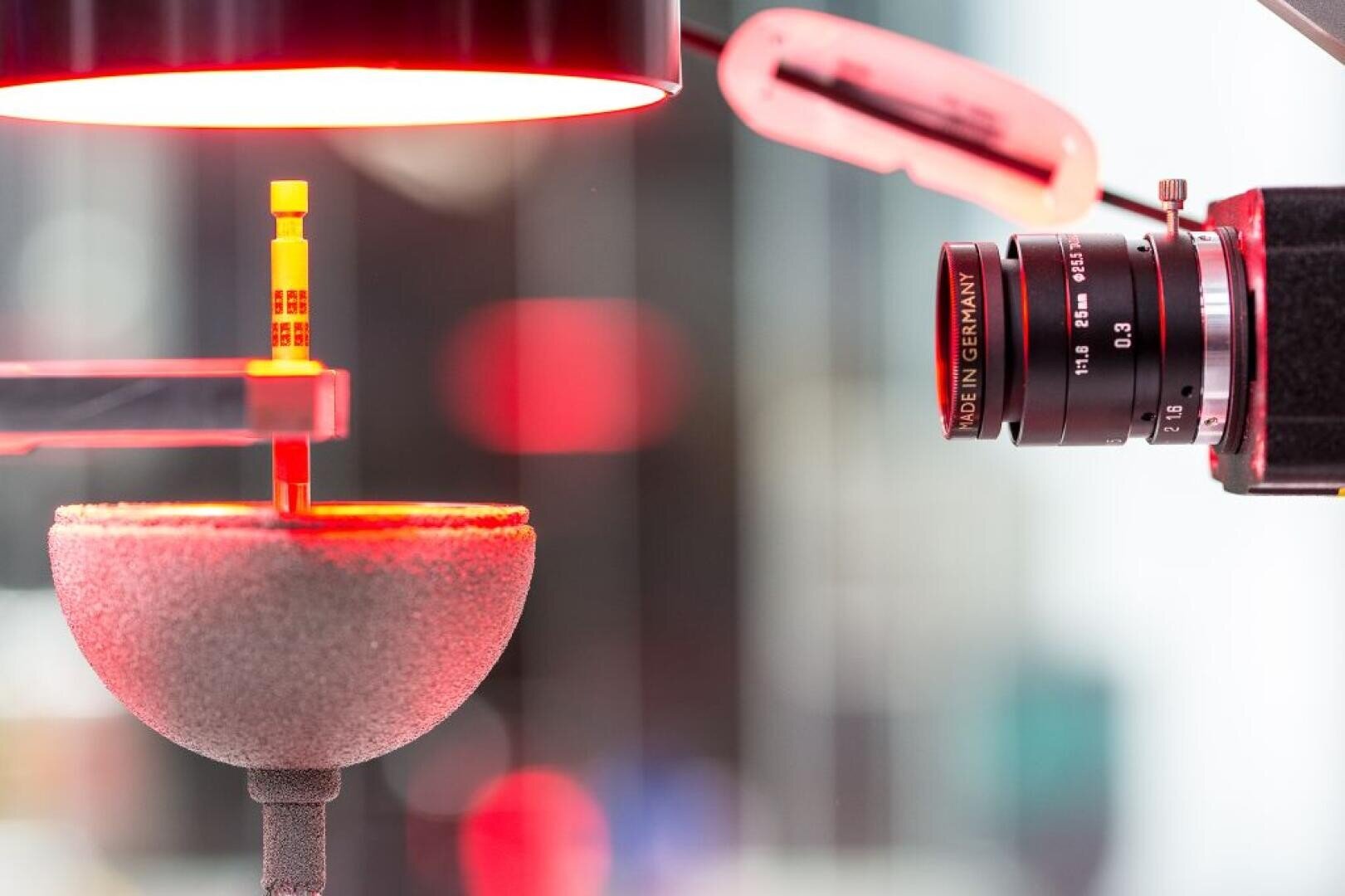 Nahaufnahme eines Kameraobjektivs, das ein kleines gelbes Objekt unter rotem Licht fokussiert, das sich über einer runden, strukturierten Plattform befindet. Der Aufbau scheint Teil eines wissenschaftlichen oder industriellen Prüfverfahrens zu sein.