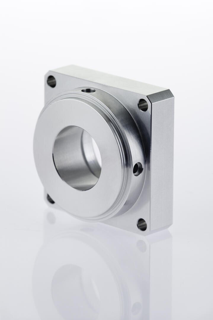 A machined metal component with a large central circular hole and four smaller corner holes, featuring a square shape and smooth metallic finish, is shown standing upright against a white background.