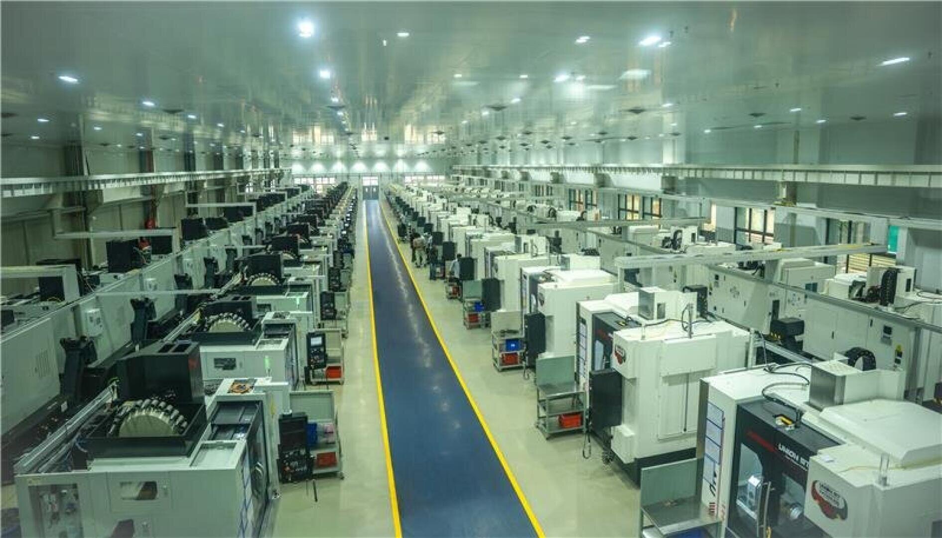 A large, brightly lit factory floor with rows of industrial CNC machines on both sides of a central blue walkway, creating a symmetrical, organized manufacturing environment.