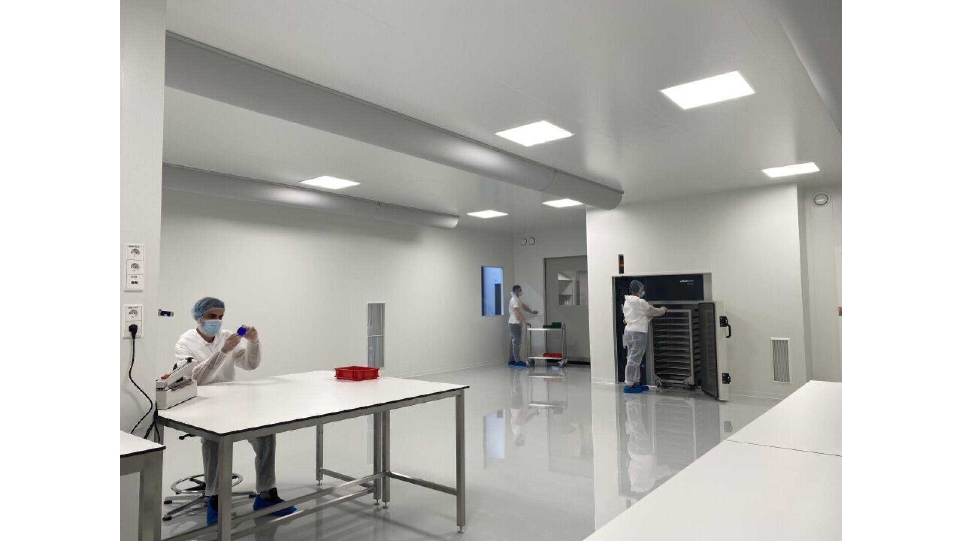 Three people wearing full protective gear work in a clean, bright laboratory room with white walls and floors. One person is at a table handling lab equipment, while two others work near large machines in the background.